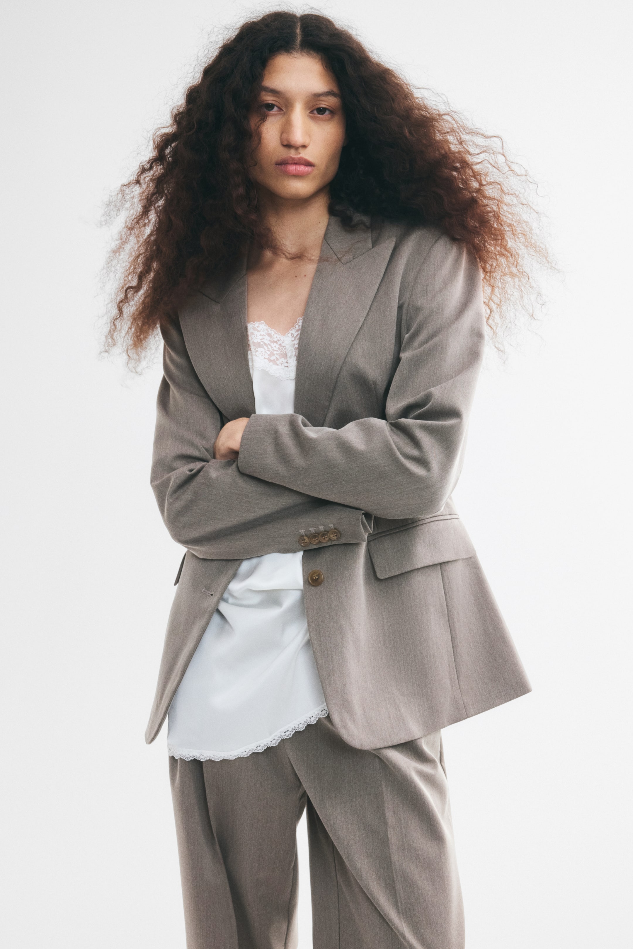 Größeres Bild ansehen: Ein langer, einreihiger Twill-Blazer in Taupe wird vom Model mit verschränkten Armen getragen und über einem weißen Camisole mit Spitzenbesatz sowie einer passenden Hose kombiniert. Der Oversize-Blazer verfügt über steigende Revers, strukturierte Schultern, geknöpfte Manschetten und Pattentaschen vorne.