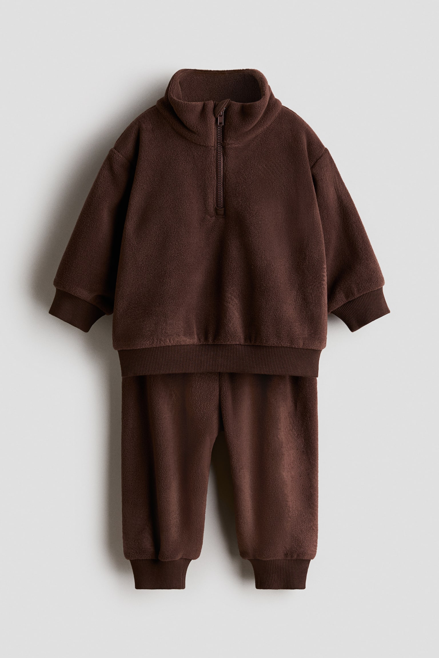 A dark brown fleece set is displayed from the front, consisting of a long-sleeved sweater and matching pants. The sweater features a stand-up collar with a half-zip closure, dropped shoulders, and ribbed trim, while the pants have ribbed cuffs at the ankles.