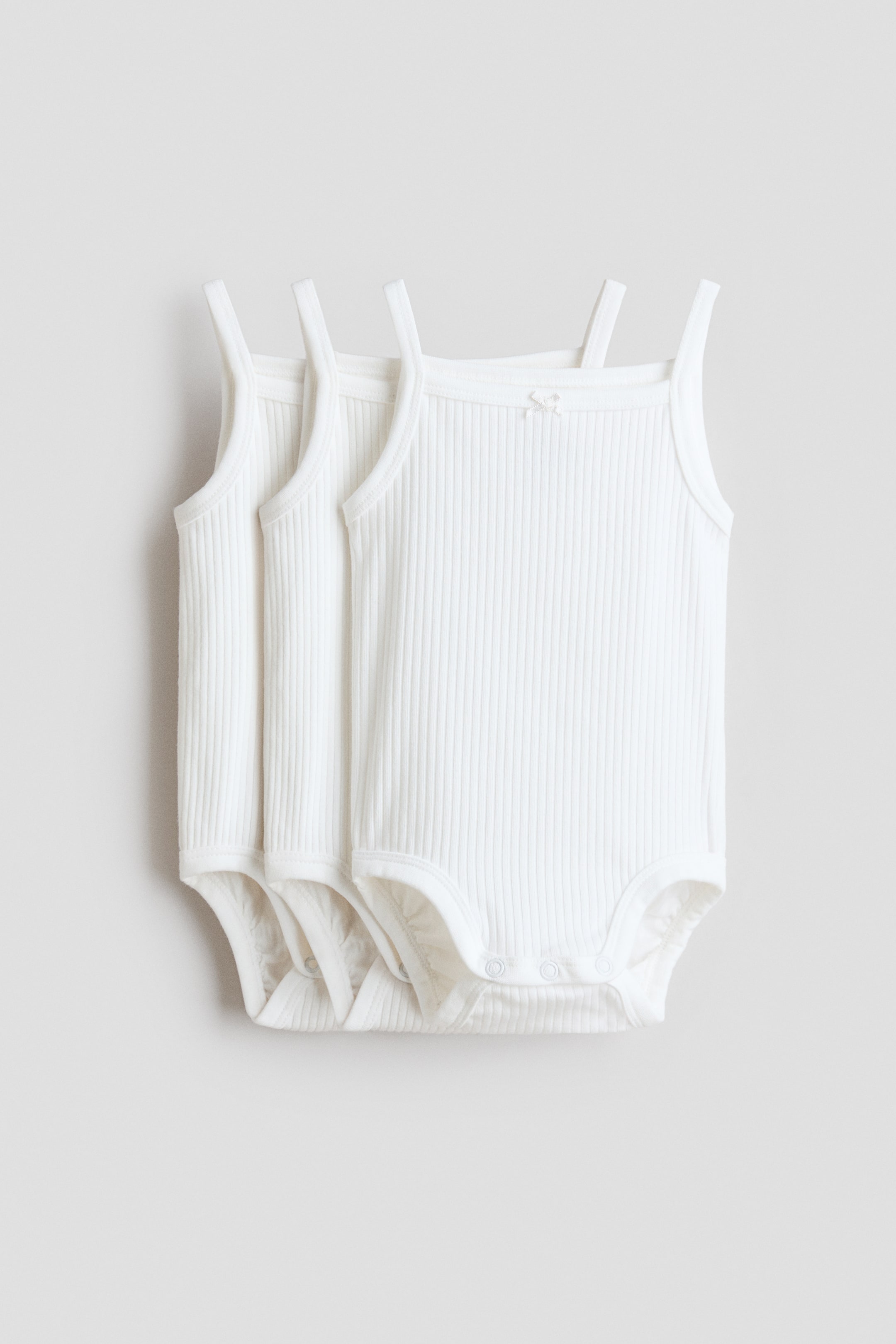 View larger image: Three white sleeveless bodysuits are showcasing a ribbed cotton jersey texture. The front of each bodysuit is visible, featuring slender shoulder straps, a square neckline, press-studs at the crotch, and a small decorative bow on the neckline of the front bodysuit.