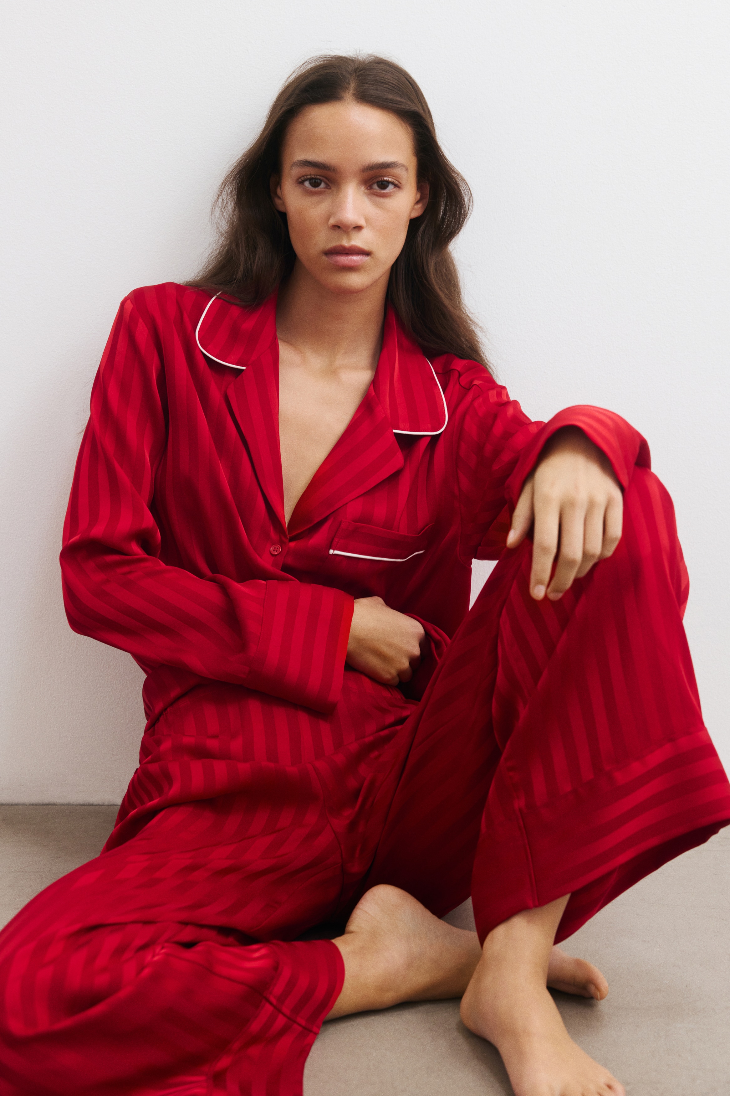A set of red textured satin pyjamas is shown from the front, featuring a long-sleeve shirt and matching straight-leg pants with a vertical stripe design. The shirt features a resort collar, a button-down front, and a chest patch pocket, with white piping accenting the collar and pocket edges.