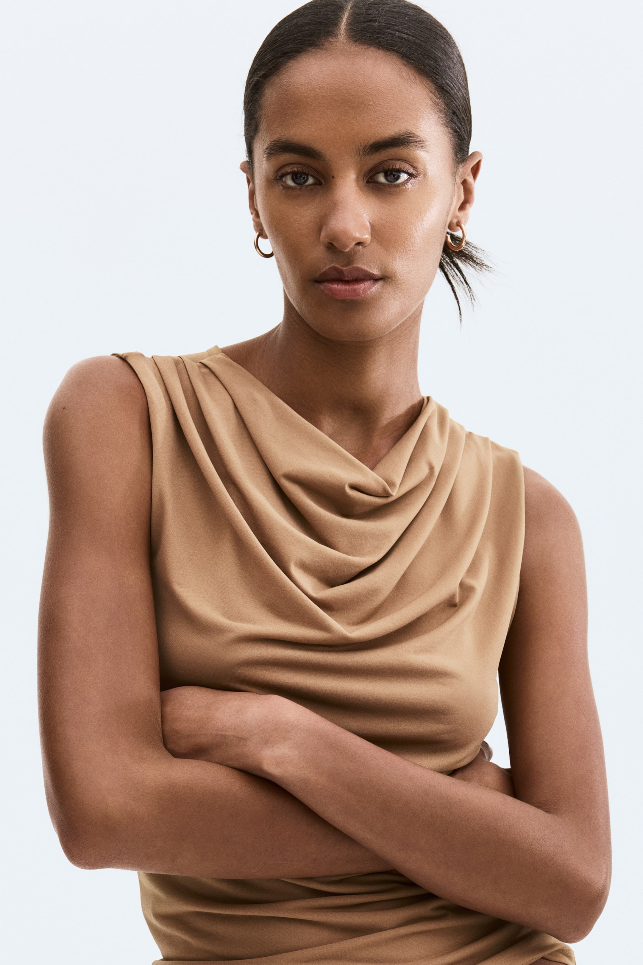 Xem ảnh lớn hơn: Áo không tay màu nâu nhạt có cổ cowl draped. Chất liệu vải mềm tạo nên những nếp gấp và xếp nếp quanh cổ áo.