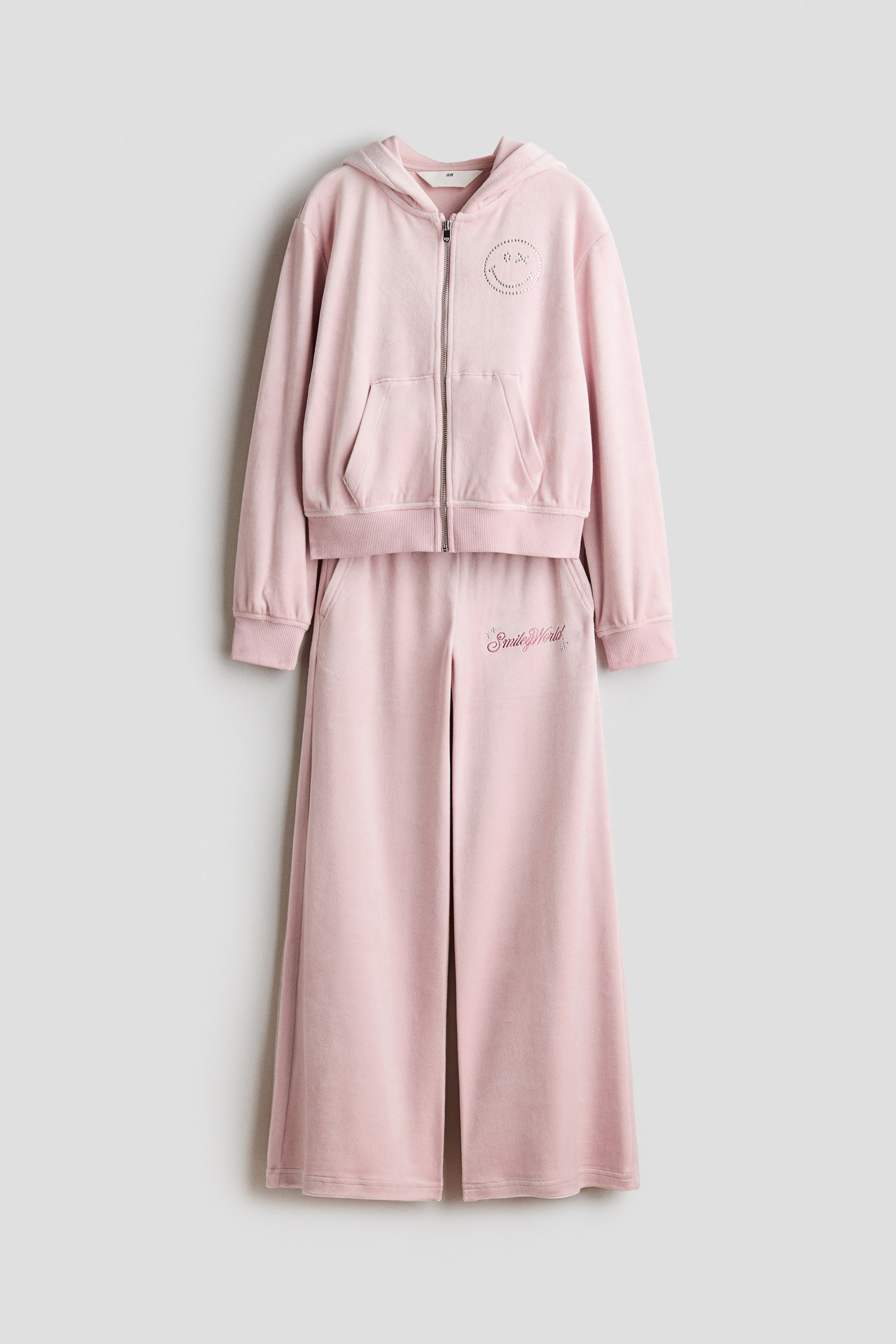 View larger image: A pink velour tracksuit is displayed, featuring a front-facing full-zip hooded jacket with a studded smiley face design on the chest, ribbed cuffs, and a ribbed hem. The matching wide-leg trousers feature "Smiley World" embroidery on the left thigh.