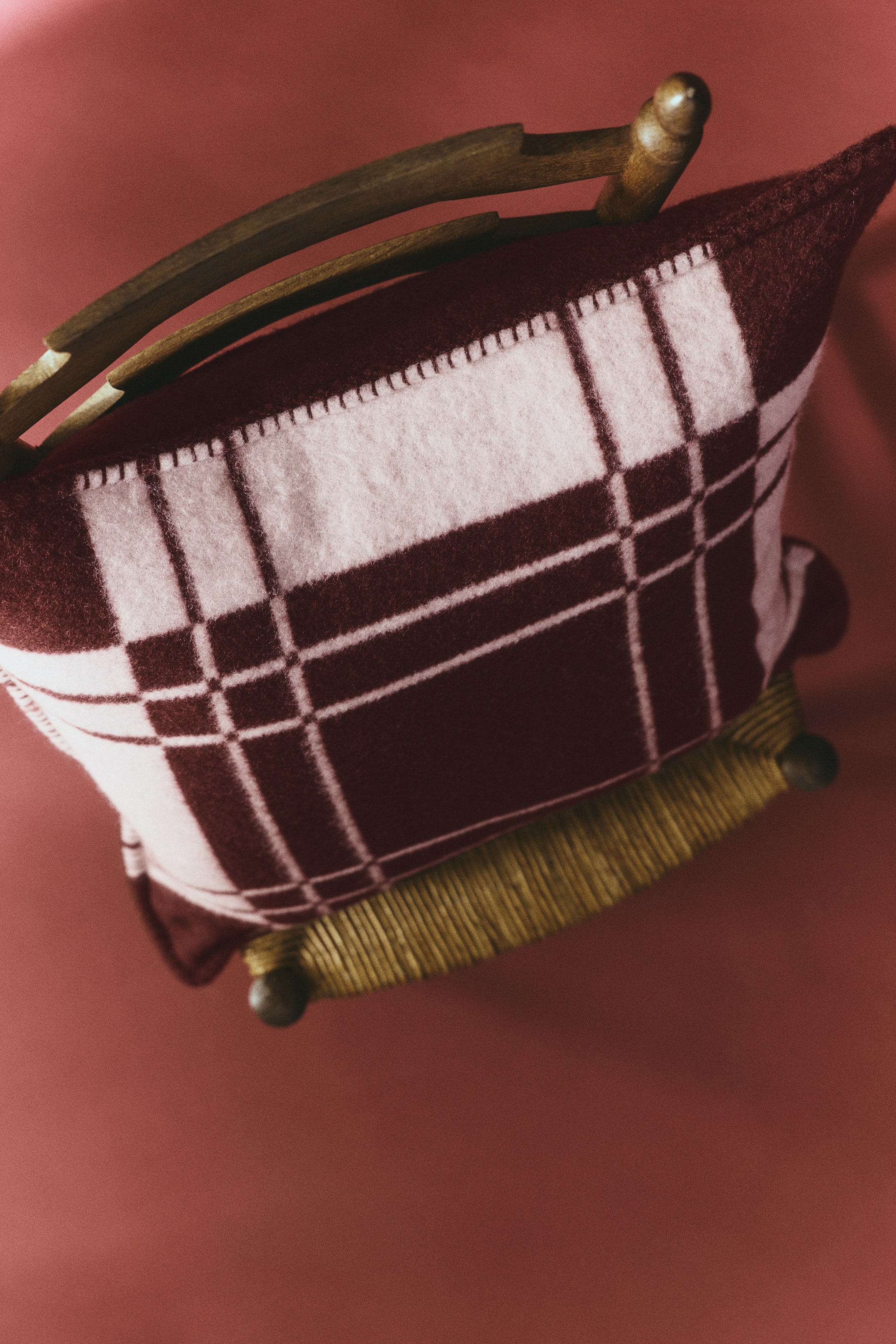 View larger image: A cushion cover in a soft, brushed wool-blend weave featuring a burgundy and light pink checked pattern rests on a wooden chair. The cushion cover displays blanket-stitched detailing along its edges, highlighting its cozy texture from a high-angle view.