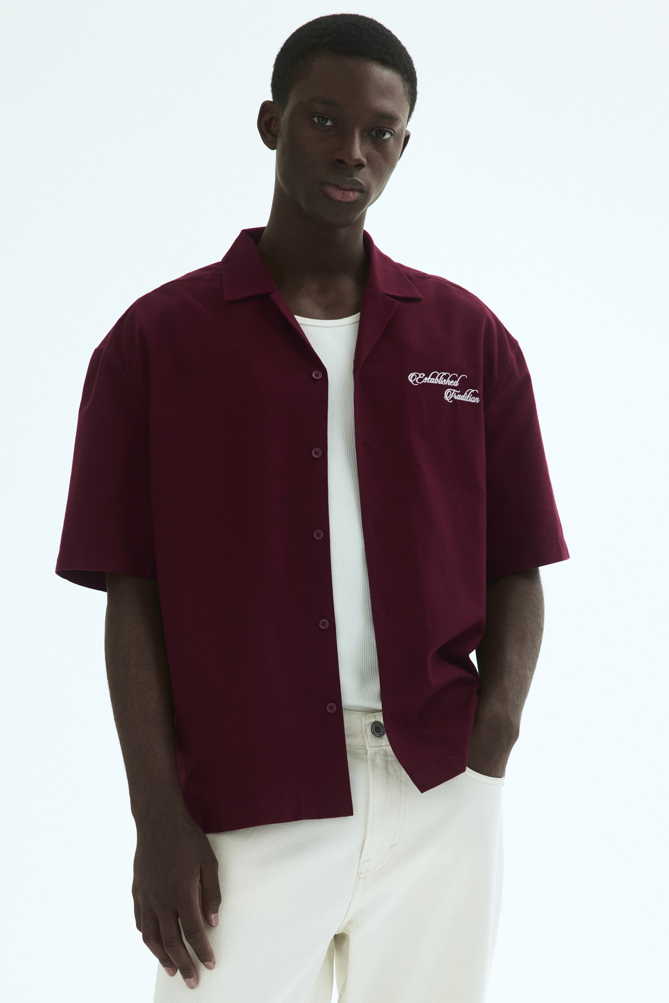 View larger image: The burgundy short-sleeved shirt showcases a resort collar and a relaxed fit, facing forward. White embroidered text reading 'Established Tradition' is visible on the left chest, and the shirt is worn open over a white ribbed tank top.