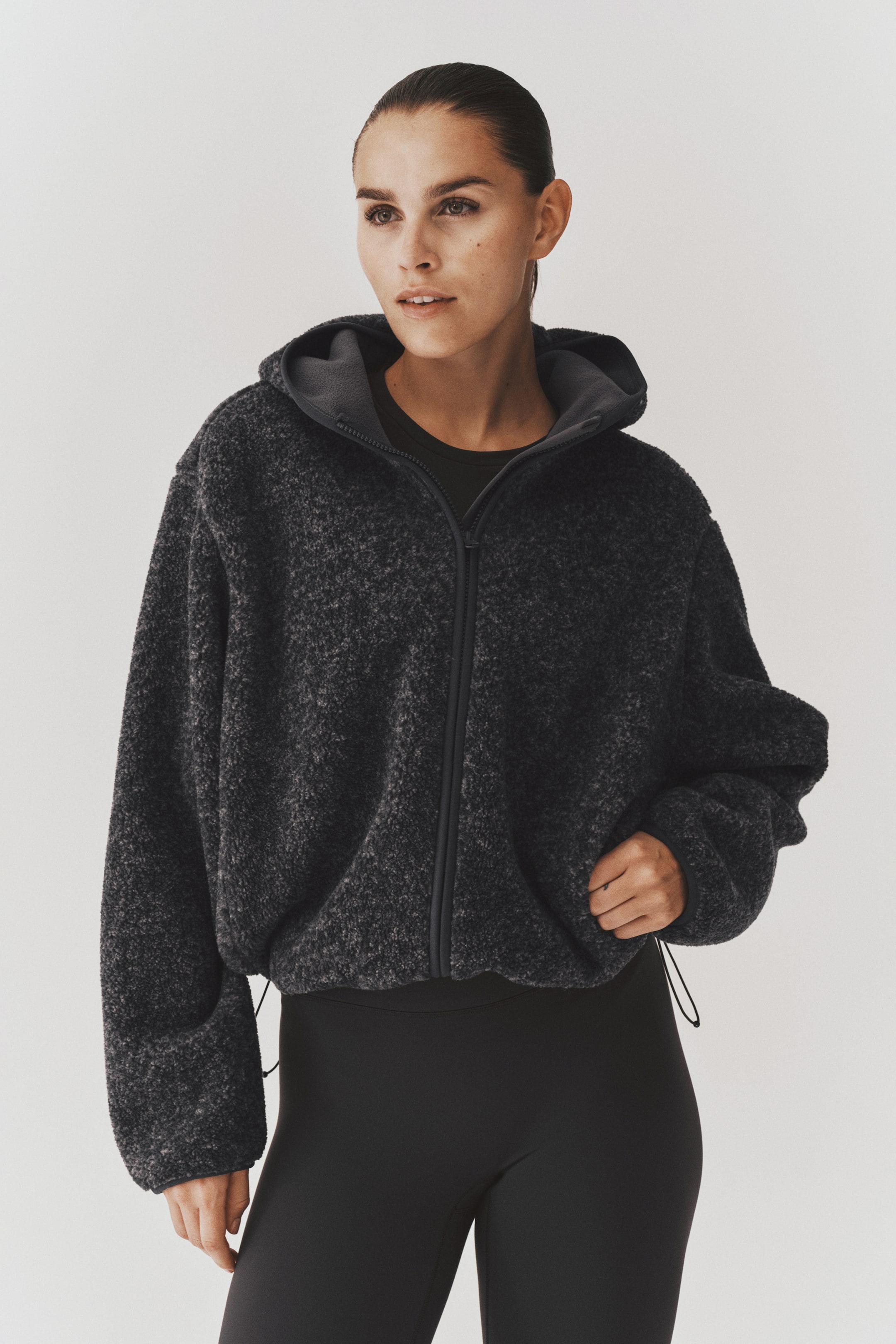 View larger image: A dark grey hooded teddy fleece jacket is being worn by the model over a black top and leggings. The loose-fit jacket features a front zip, long sleeves with elasticated cuffs, and an adjustable drawstring hem with cords hanging at the sides.
