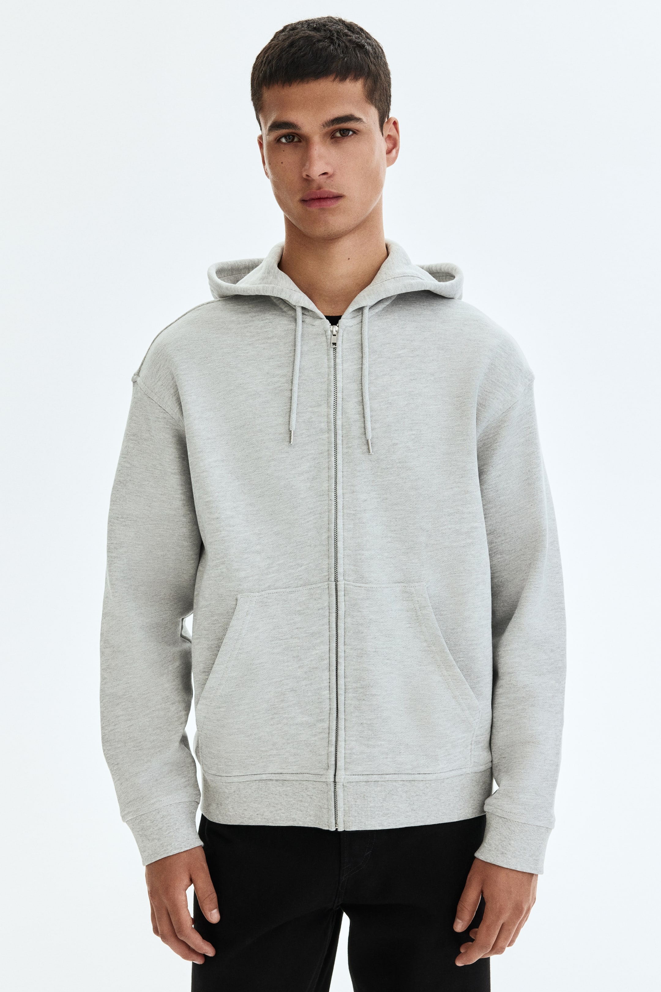 View larger image: The model is showcasing a grey zip-through hoodie. It boasts a partially zipped front, a hood, and front pockets. The design includes dropped shoulders, long sleeves with ribbed cuffs, and a ribbed hem.