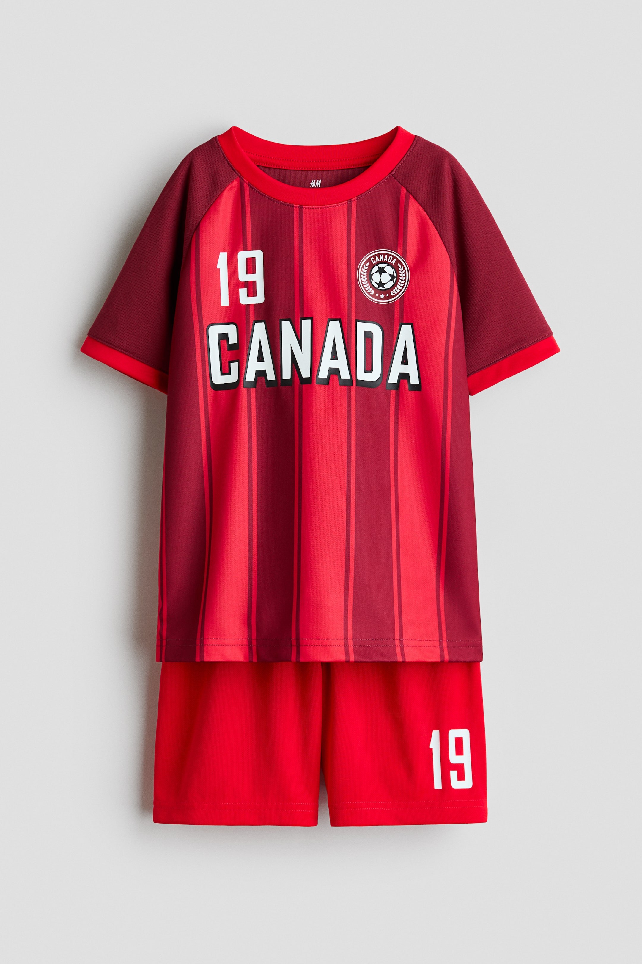 View larger image: Printed soccer set in soft mesh fabric featuring a short-sleeved top and a pair of shorts with an elastic, drawstring waist is shown from the front. The top showcases a red body with darker red vertical stripes and darker red raglan sleeves, displaying '19' and 'CANADA' in white on the chest, along with a circular soccer emblem. The matching red shorts display '19' in white on the right leg.
