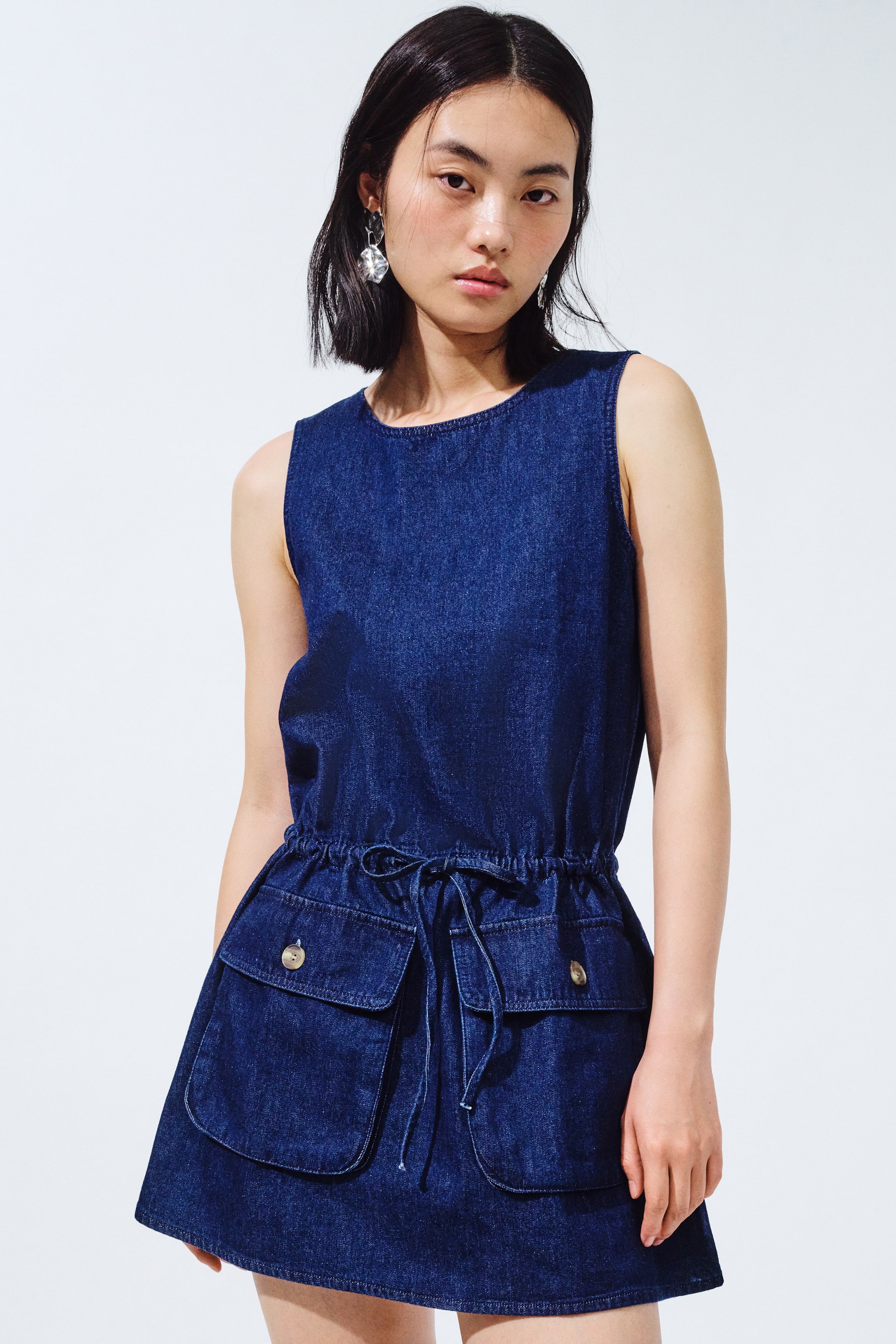 Xem ảnh lớn hơn: Chiếc váy denim màu xanh đậm có thiết kế cổ tròn và không tay. Nó được thắt eo bằng dây rút, và hai túi vá lớn phía trước với nắp có nút là điểm nhấn cho phần chân váy.