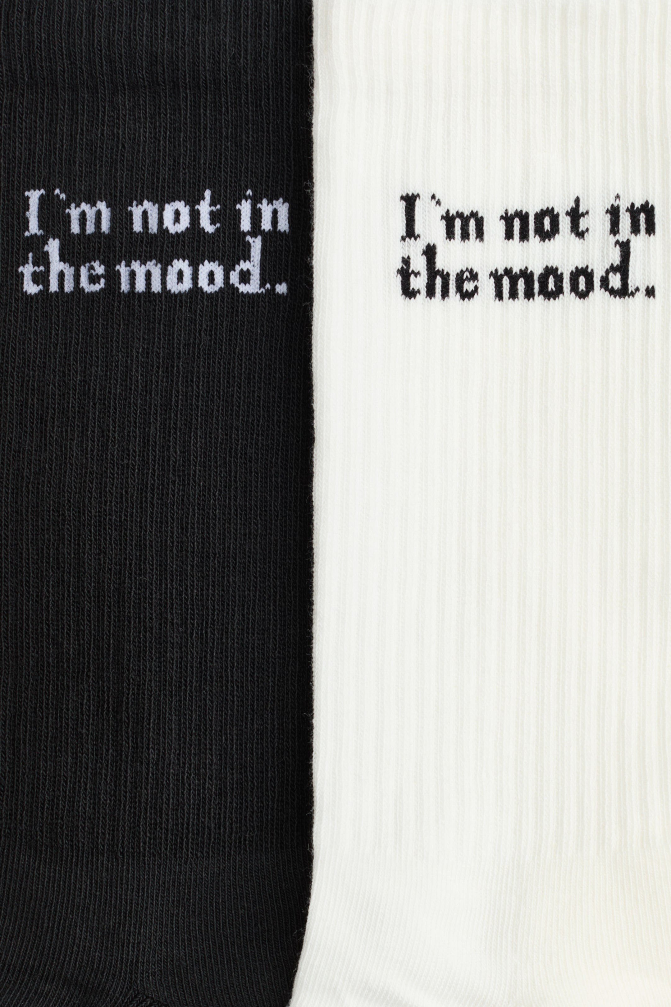 Zobacz większy obraz: Dwie skarpetki, jedna czarna i jedna biała, są wyeksponowane pionowo obok siebie. Obie mają wysokie, prążkowane cholewki i prezentują kontrastowy napis: „Nie mam humoru”.