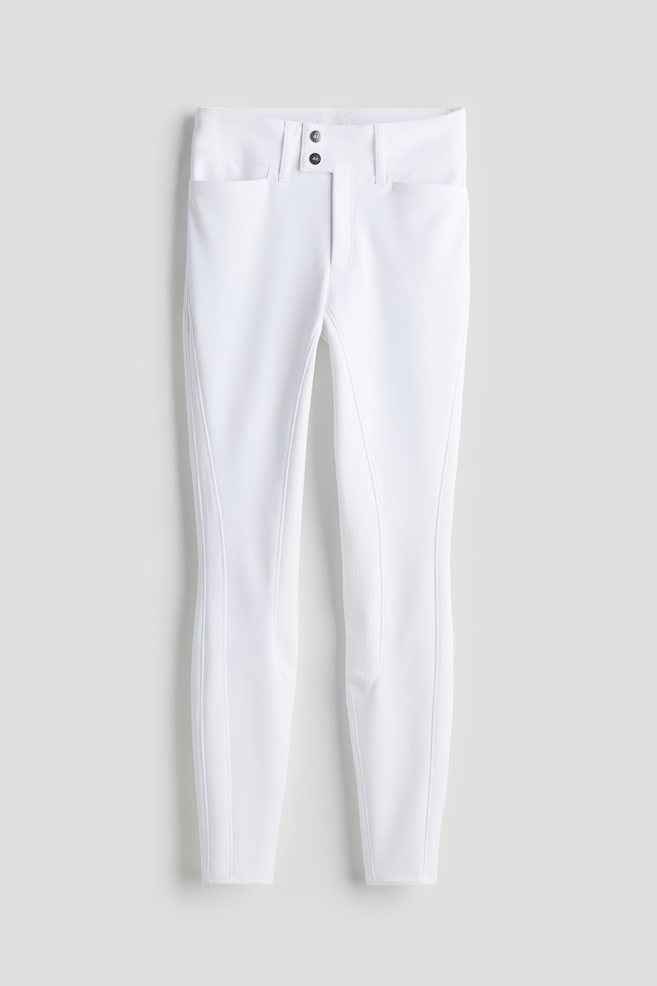 White breeches are displayed facing FRONT, featuring a high waist with a zip fly and an extended waistband secured by two dark decorative buttons, alongside two open, slanted front pockets. Textured panels, indicative of silicone grip, are visible on the inner legs, with narrow, cleanly finished hems completing the design. - 1