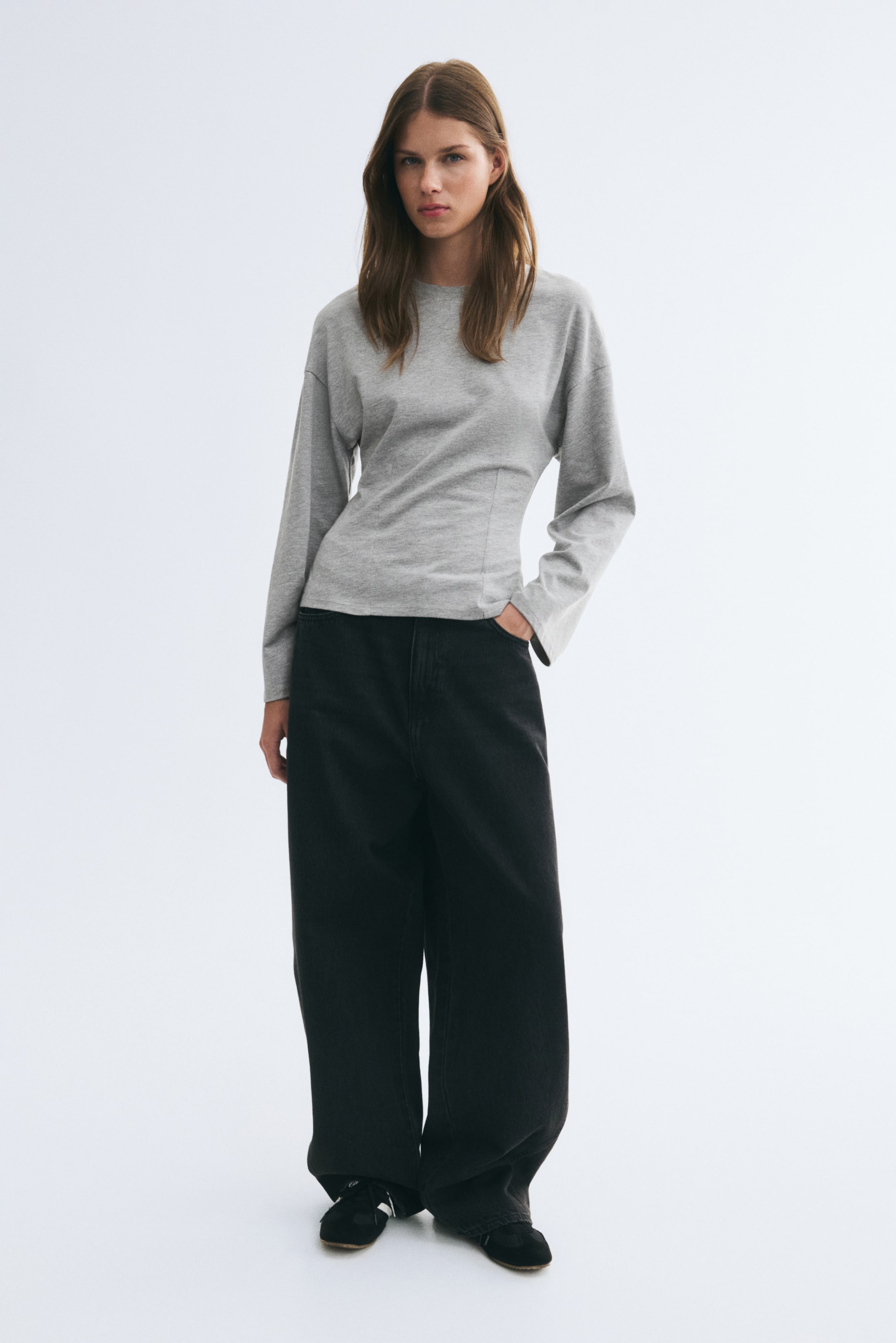 Größeres Bild ansehen: Das Model trägt ein hellgraues Langarmshirt mit Rundhalsausschnitt, überschnittenen Schultern und einer figurbetonten, strukturierten Taille. Das Oberteil ist mit einer schwarzen Wide-Leg-Jeans kombiniert, während das Model eine Hand lässig in der vorderen Tasche ablegt.