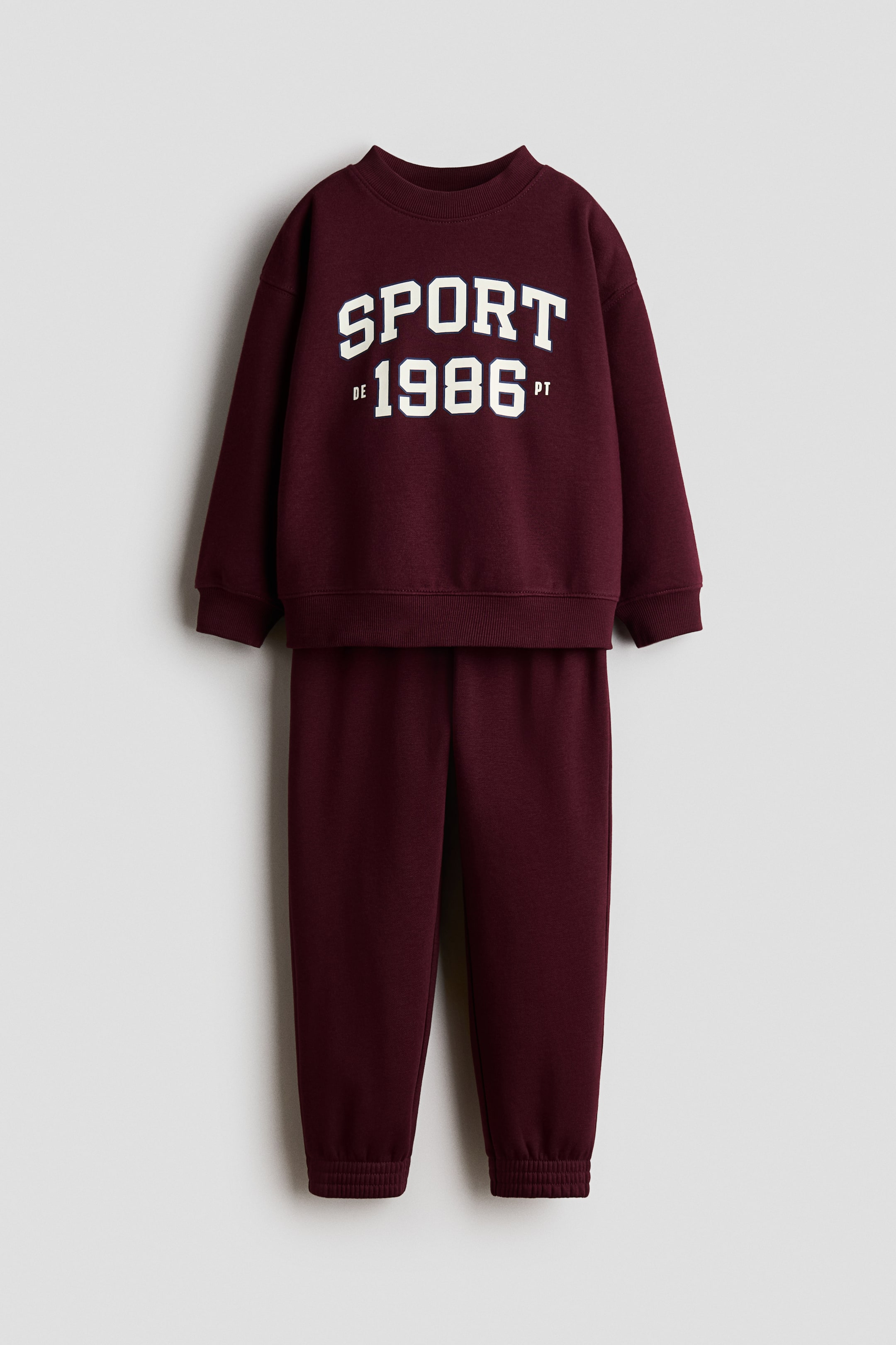 Visa större bild: Detta vinröda träningsoverallset innehåller en crewneck-sweatshirt med långa ärmar, ribbade muddar, en ribbad nederkant och en vit "SPORT DE 1986 PT"-grafik på framsidan. Matchande träningsbyxor kompletterar setet med ribbade ankelmuddar.