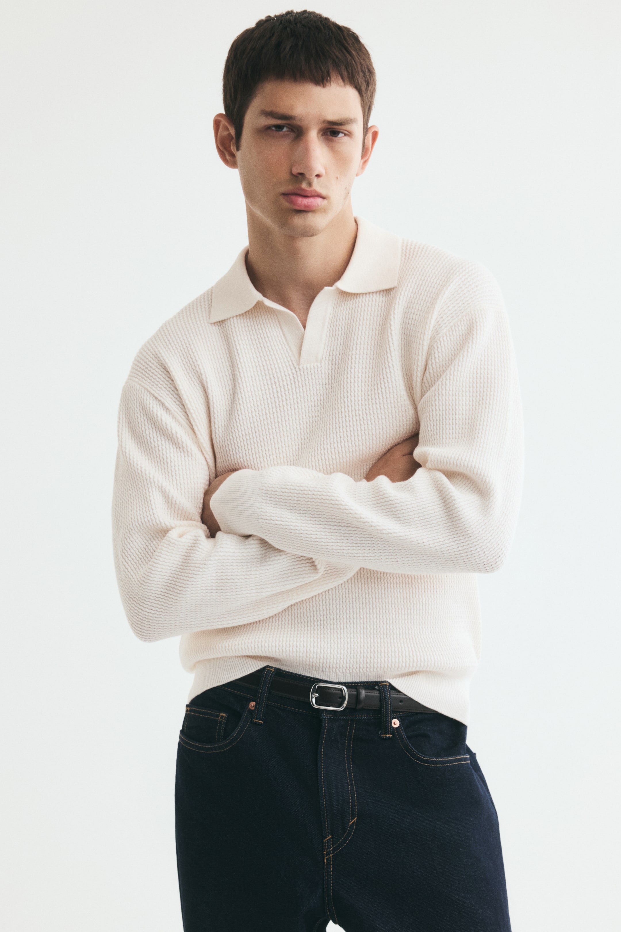 View larger image: A textured-knit, long-sleeve polo shirt in an off-white cotton blend is worn by the model with arms crossed. The relaxed-fit shirt features an open collar and ribbed cuffs, and is styled tucked into dark blue jeans secured with a black belt.