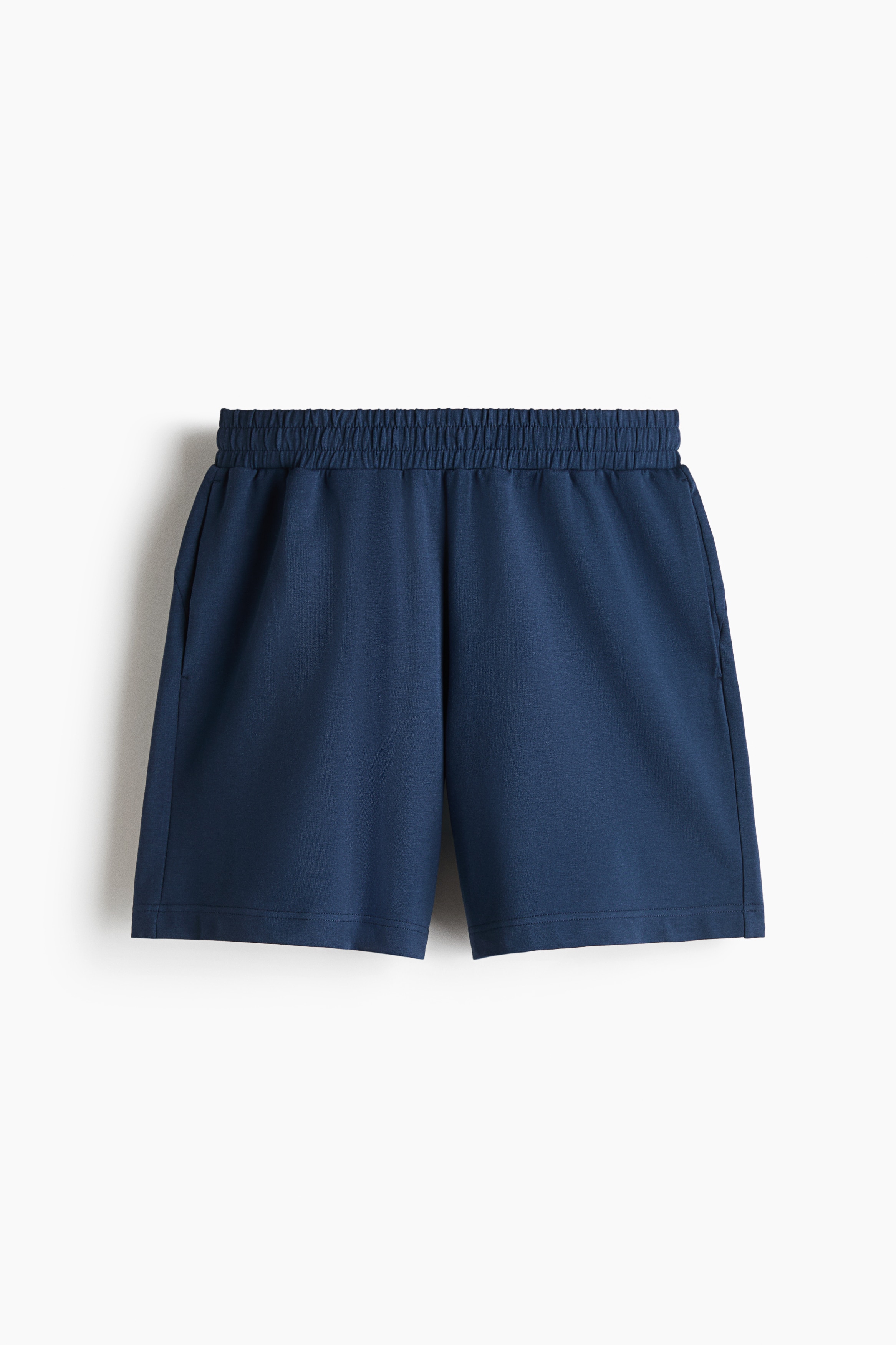 Blue sweat shorts featuring an elasticated waistband and a regular fit are being worn by the model, who is standing with one hand resting inside a side pocket. The shorts are styled with a matching blue, textured long-sleeve top.