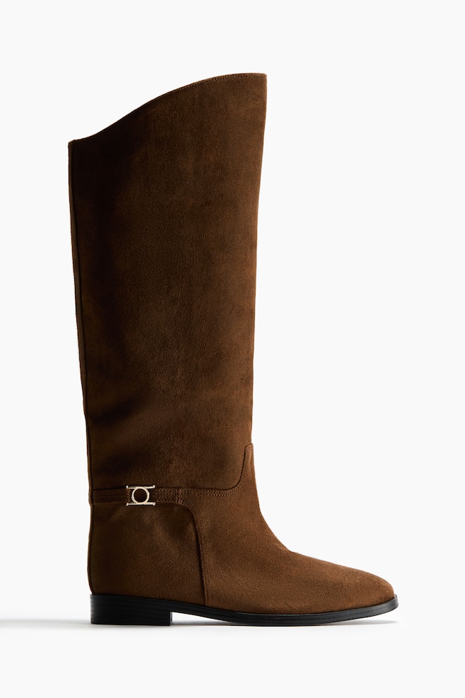 A brown suede-effect boot features a tall shaft with a subtly curved top edge. A decorative gold-toned, circular buckle adorns a strap around the ankle, and a flat black sole provides a low block heel. - 1
