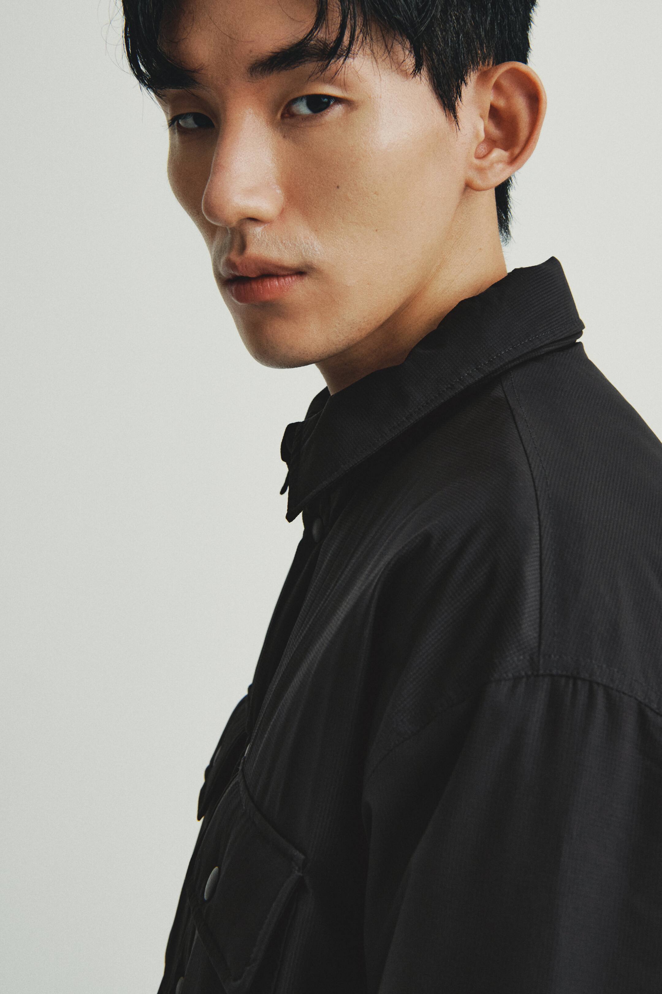 이미지 확대 보기: 블랙 셰킷은 클래식한 뾰족한 칼라와 미세한 텍스처 짜임이 돋보이는 측면-앞 각도에서 보여집니다. 착용자의 왼쪽 가슴에는 스냅 잠금 장치가 있는 큰 플랩 포켓이 있으며, 여유로운 핏을 선보이고 있습니다.