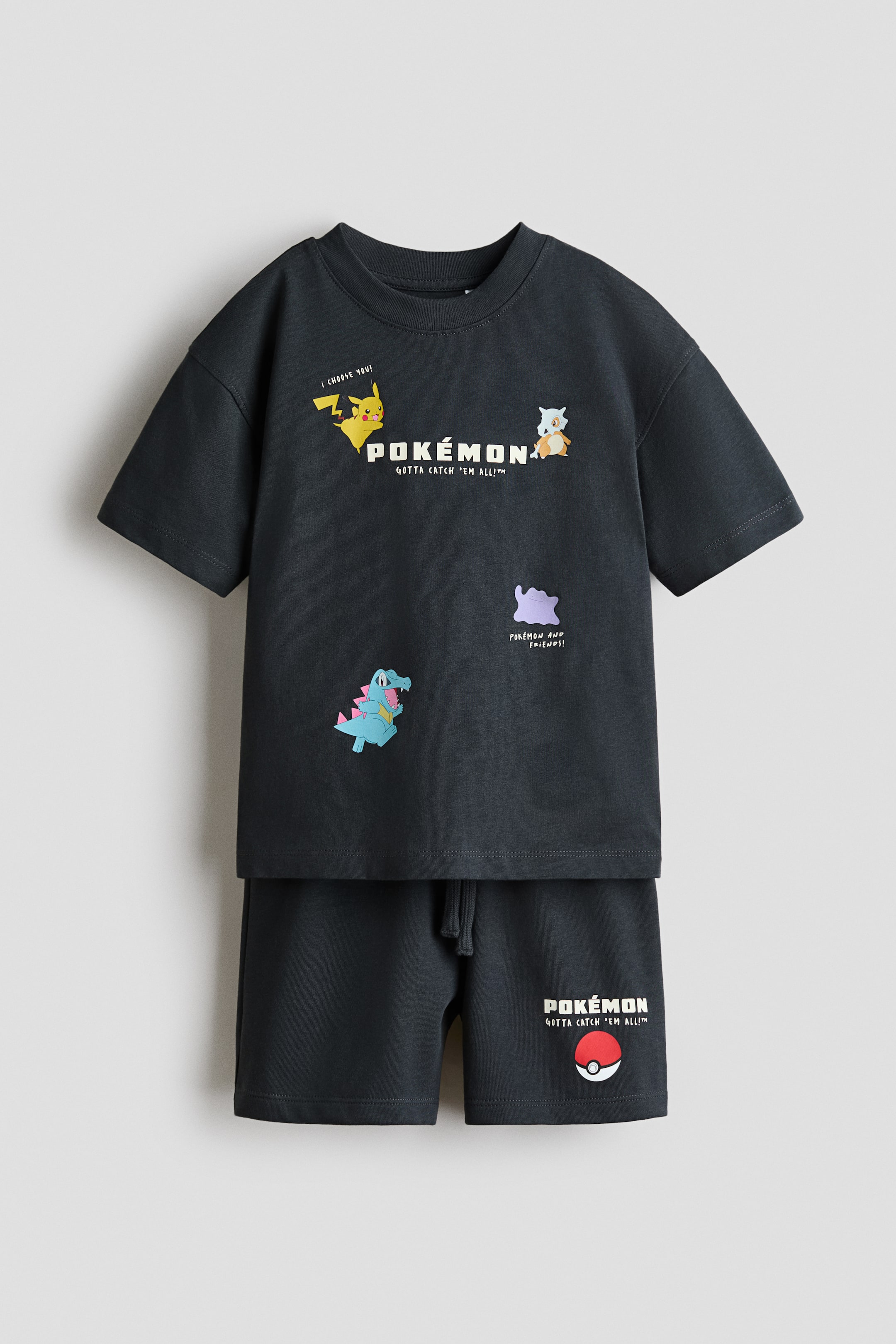 Ver imagem maior: Um conjunto de T-shirt e calções a condizer em cinzento-escuro é apresentado de frente. A T-shirt de jersey de algodão apresenta ombros descaídos, mangas largas, uma gola canelada e várias estampas de personagens e textos de Pokémon, enquanto os calções leves de moletom apresentam uma cintura com cordão e um motivo de Poké Ball com o texto 'Pokémon Gotta Catch 'Em All!?'.