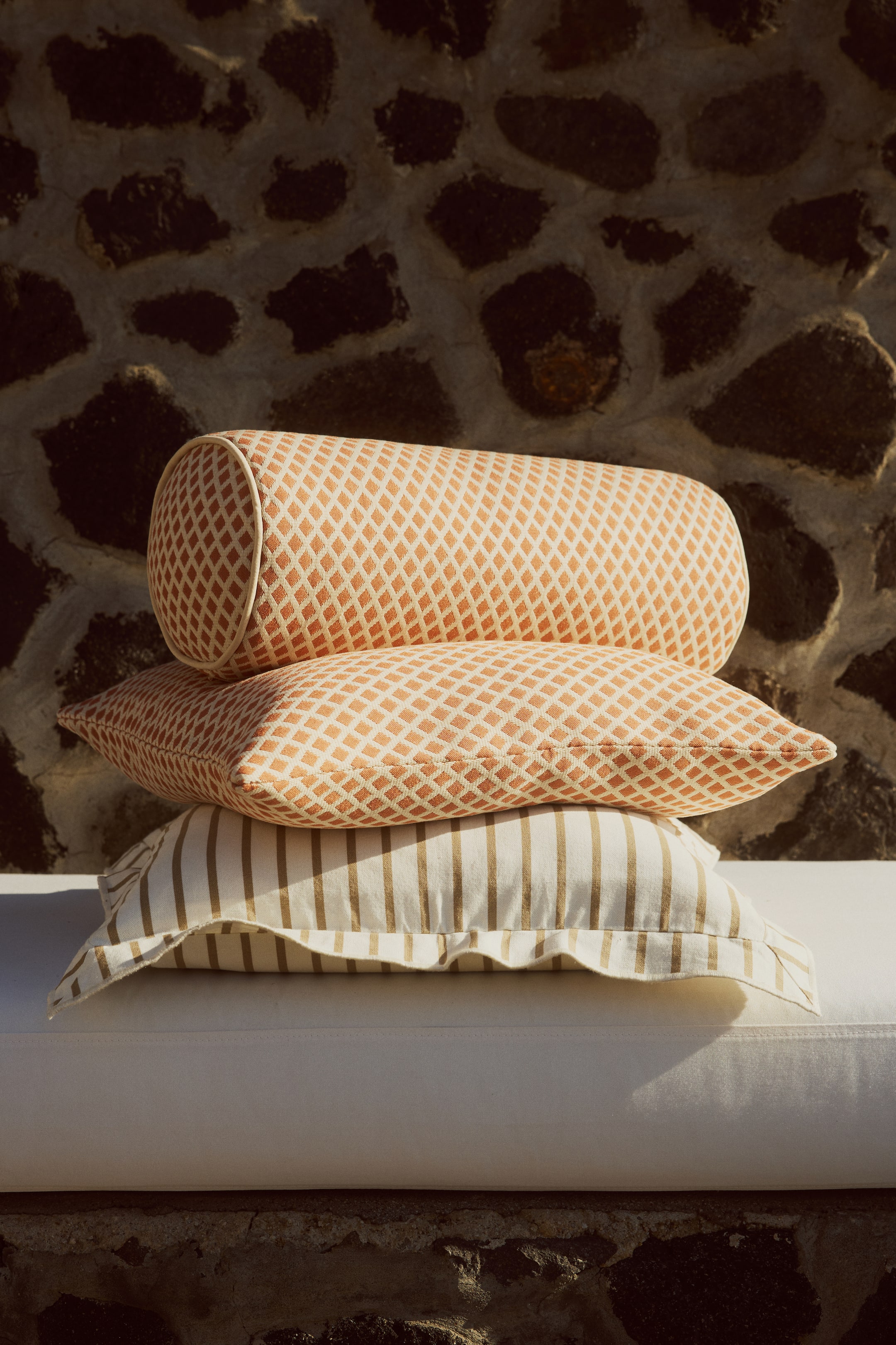 View larger image: A cylindrical bolster pillow is lying horizontally atop a rectangular throw pillow, both showcasing an orange and off-white diamond pattern accented with off-white piping. Below them, a second rectangular throw pillow displays vertical beige and off-white stripes, completing the stack.