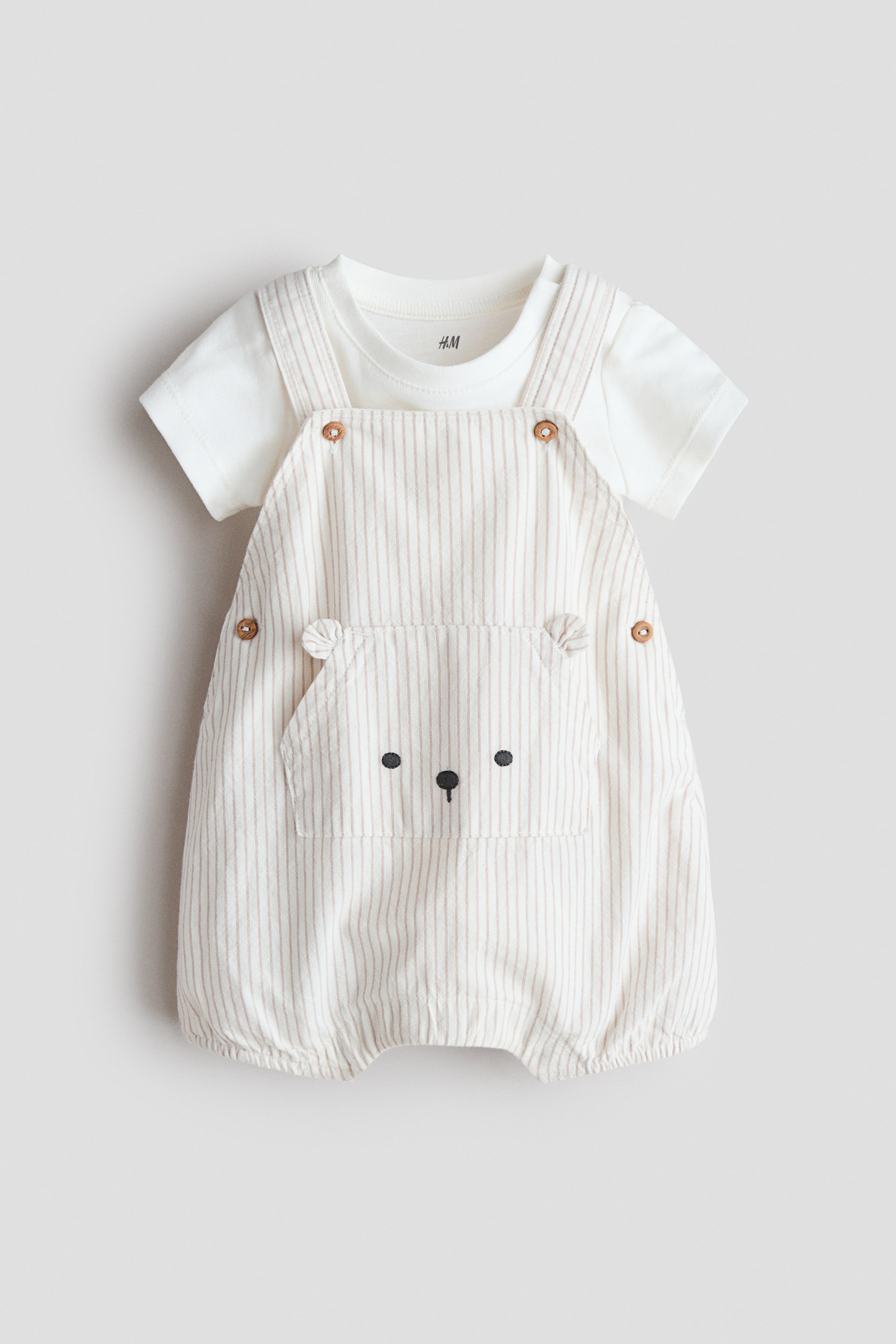 View larger image: A baby clothing set is showcased from the front, featuring a white short-sleeved bodysuit layered under vertically striped beige dungarees. The dungarees highlight a large front kangaroo pocket adorned with an embroidered bear face, adjustable buttons on the straps and sides, and elasticated leg openings.