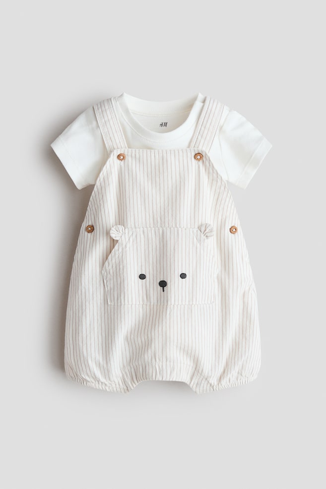 A baby clothing set is presenting from the front, featuring a white short-sleeved bodysuit layered under vertically striped beige dungarees. The dungarees are displaying a large front kangaroo pocket with an embroidered bear face, adjustable buttons on the straps and sides, and elasticated leg openings. - 1