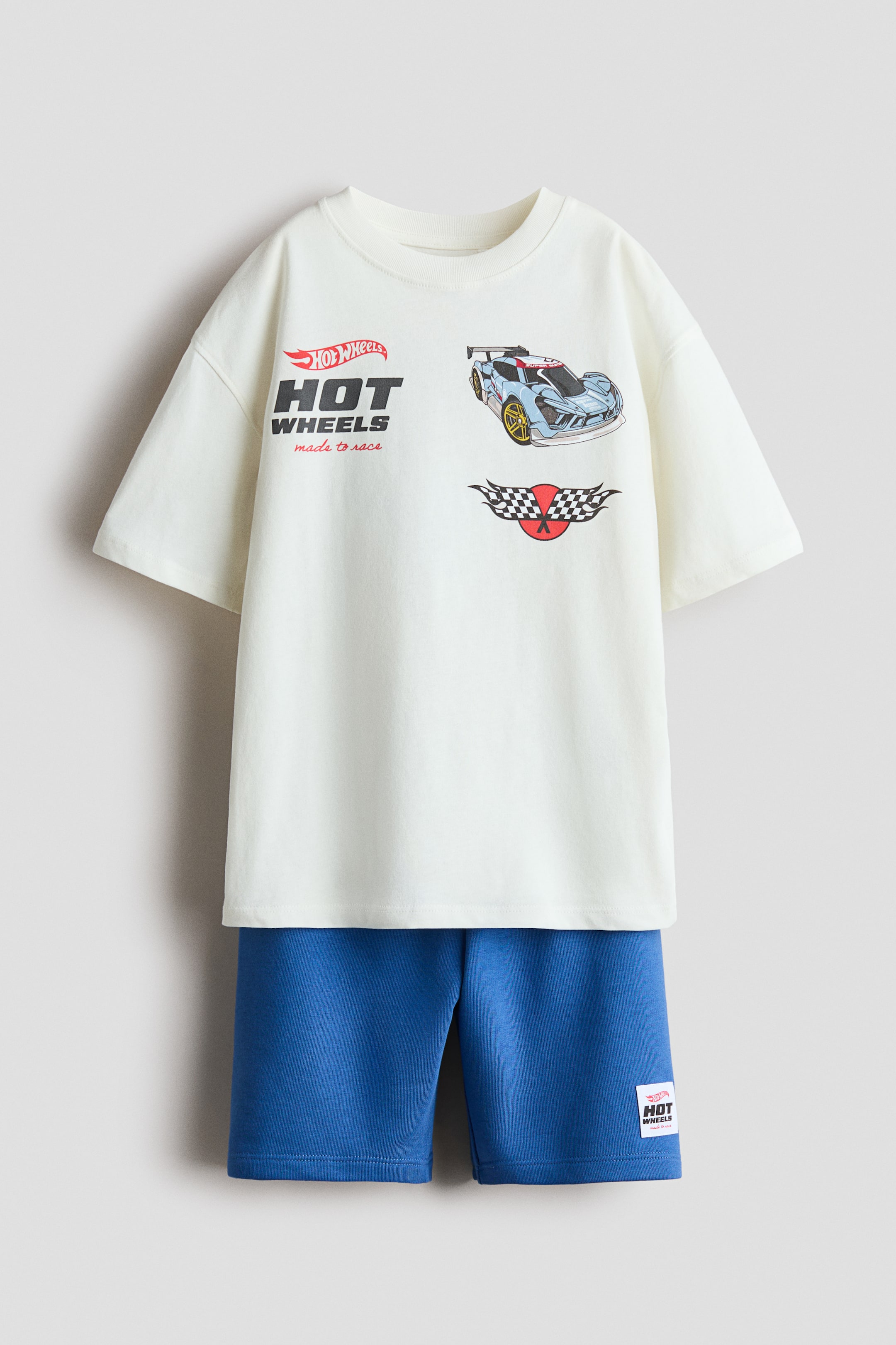 Größeres Bild ansehen: Ein passendes Set präsentiert ein cremefarbenes T-Shirt und blaue Shorts, beide von vorne gezeigt. Das cremefarbene T-Shirt ist mit überschnittenen Schultern, weiten Ärmeln und einem gerippten Halsausschnitt gestaltet und zeigt Hot Wheels Auto- und Logodrucke, während die blauen Shorts ein kleines Hot Wheels-Label auf dem rechten Bein aufweisen.