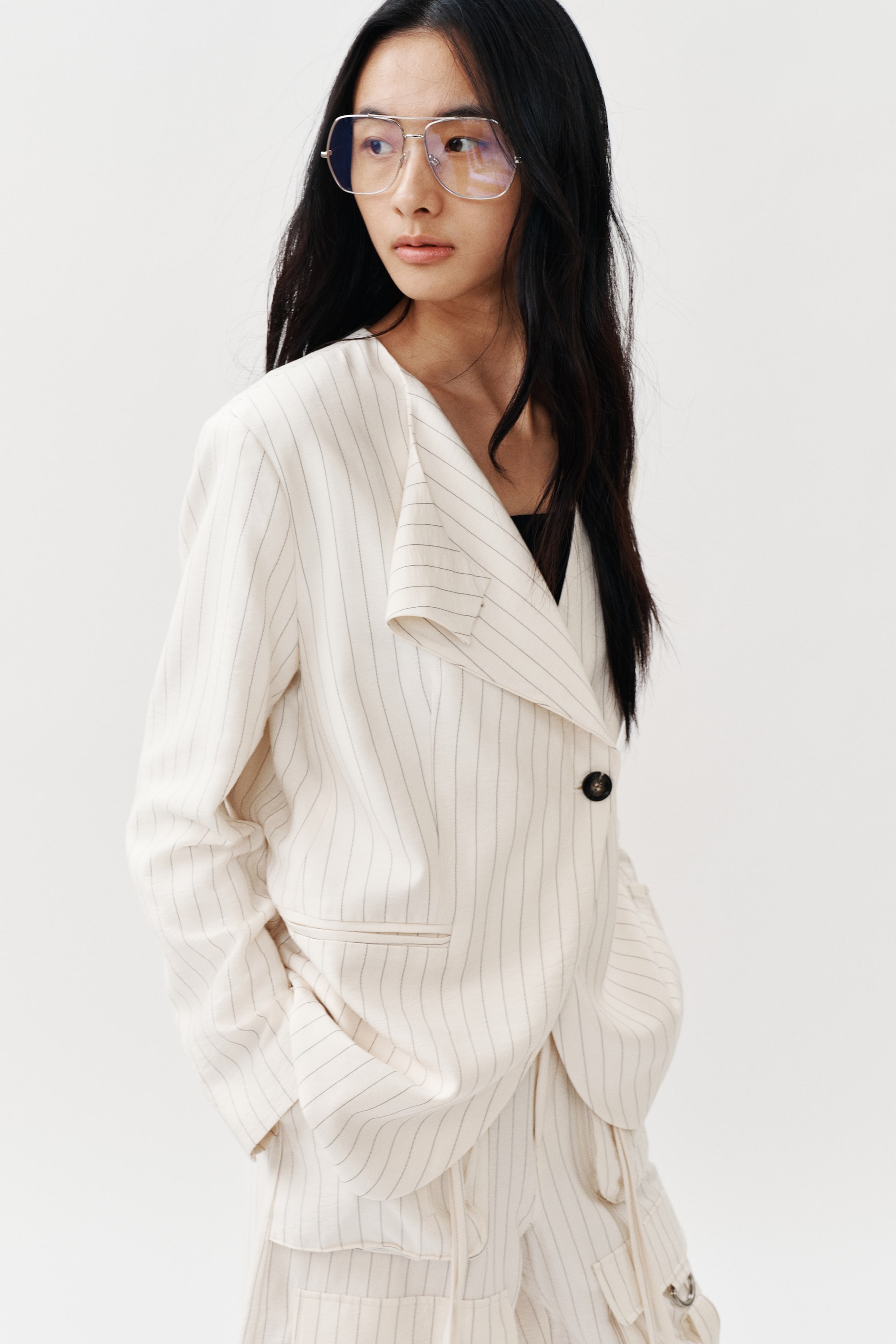 View larger image: A cream pinstriped blazer, angled slightly to the left, showcases an oversized, asymmetric lapel with a single dark button closure. It's styled over a black top and paired with matching pinstriped trousers featuring visible utility pockets.