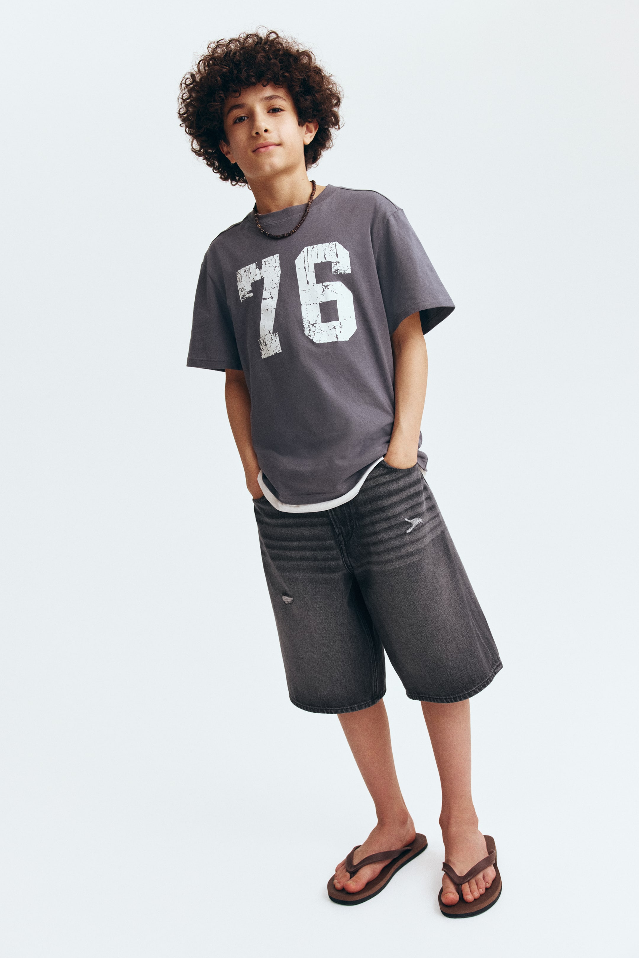 View larger image: A grey short-sleeved t-shirt na may malalaking distressed na puting numero "76" sa harap, suot ito sa ibabaw ng puting undershirt na bahagyang nakikita sa laylayan. Ipinapareha ito sa grey denim shorts na may distressed na detalye, kabilang ang faded na pahalang na mga guhit at maliit na punit sa kaliwang hita.