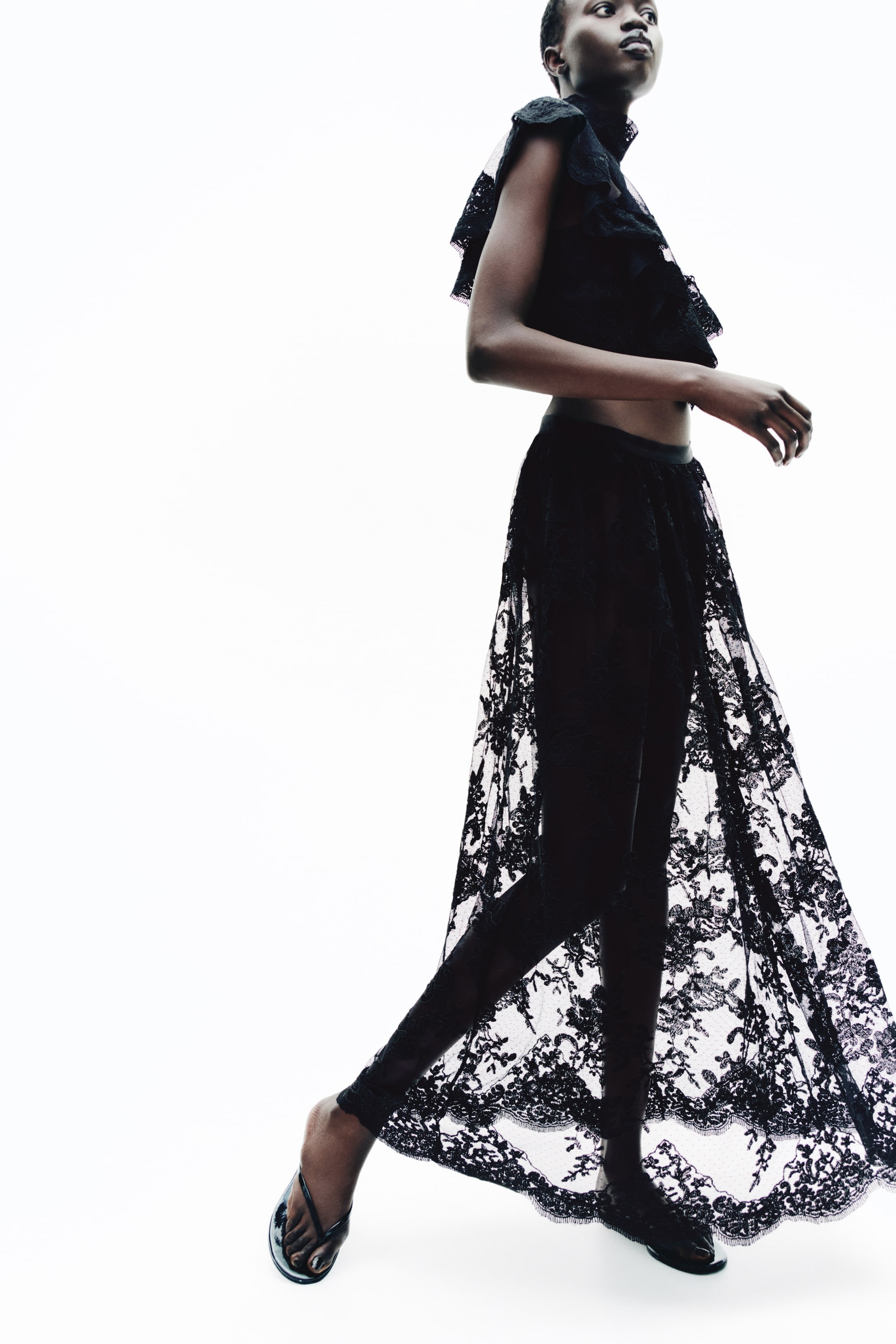 查看大圖: 模特兒穿著一件黑色花卉蕾絲長裙,寬鬆版型,展現出半透明、無內裡的垂墜感與略帶 A 字的輪廓。裙身採用波浪花邊裙擺,並搭配黑色緊身褲及同款蕾絲上衣。