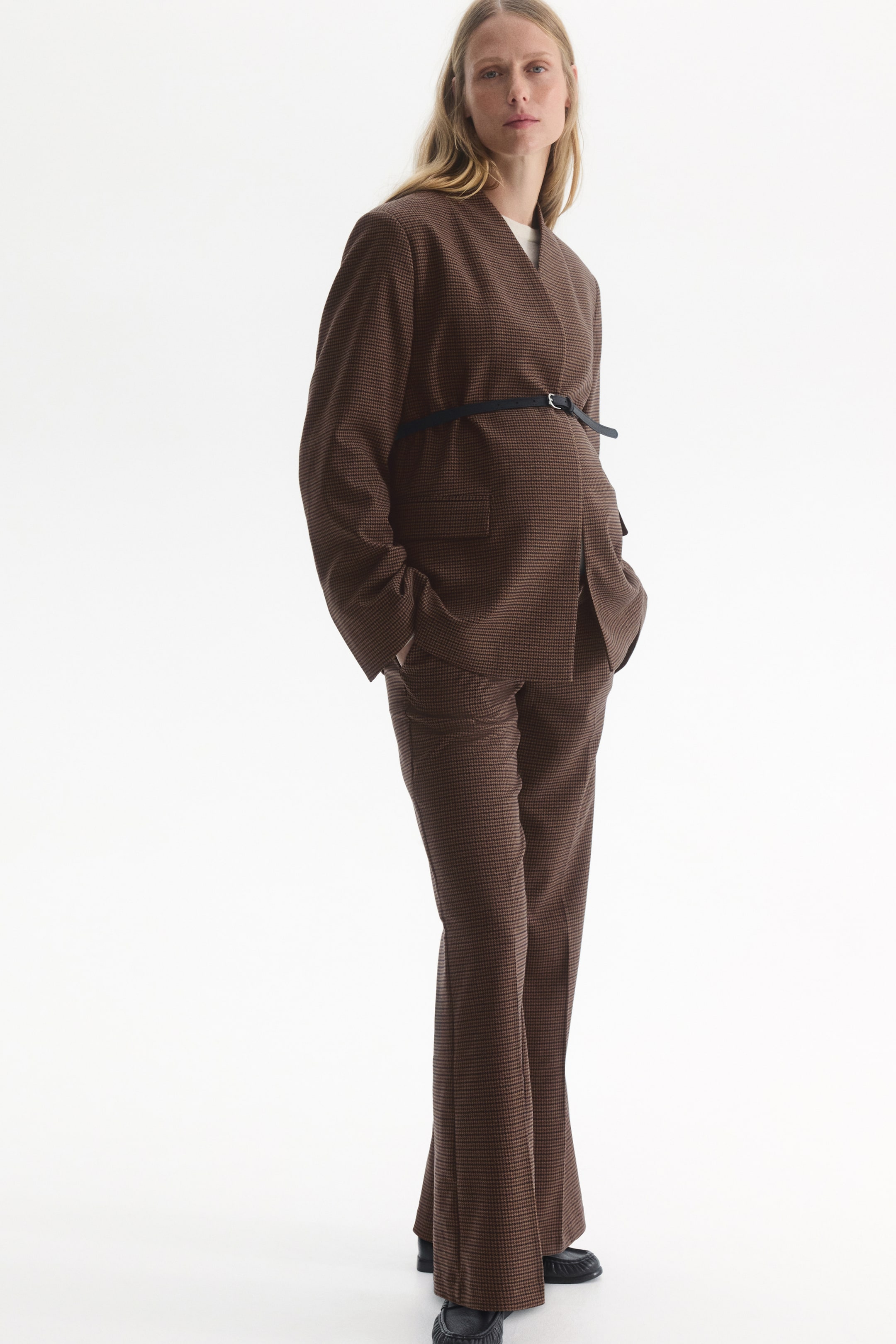 Grotere afbeelding bekijken: Bruine broek van tweedstof met patroon, met bootcut-pijpen en strakke plooien langs de voorkant, gedragen door het model. De broek is gestyled met een bijpassende blazer, vastgehouden met een dunne zwarte riem boven de taille, en zwarte loafers.