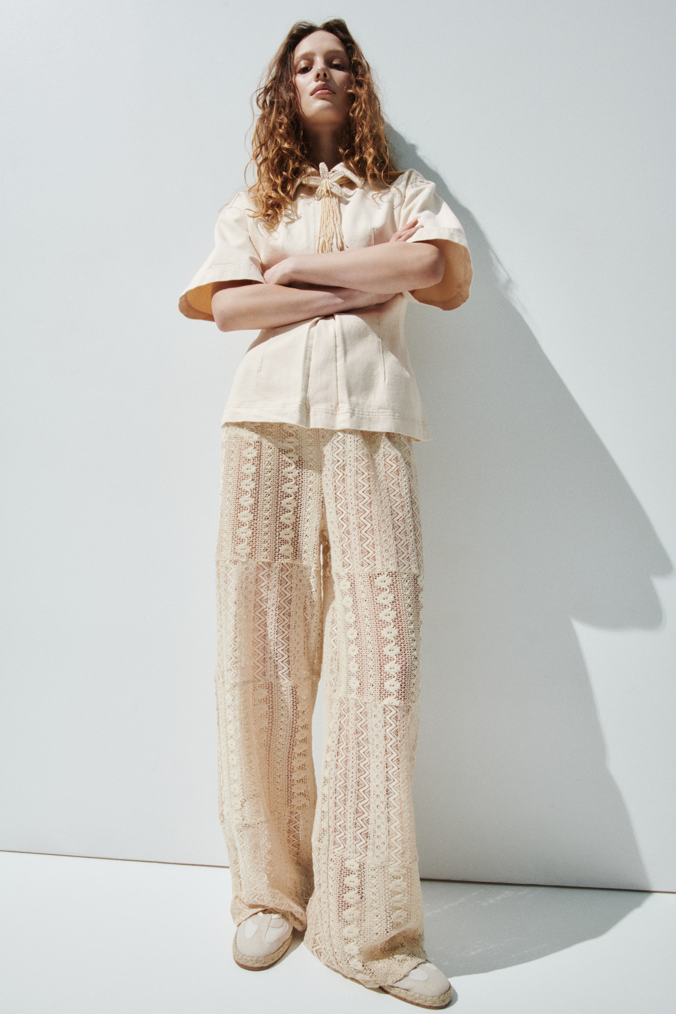 Ingrandisci l'immagine: Un top a maniche corte di colore beige chiaro e pantaloni a gamba larga sono presentati. Il top presenta un colletto, un'apertura a V con un fiocco intrecciato e nappine, e cuciture verticali. I pantaloni a gamba larga mostrano un intricato motivo in pizzo trasparente.