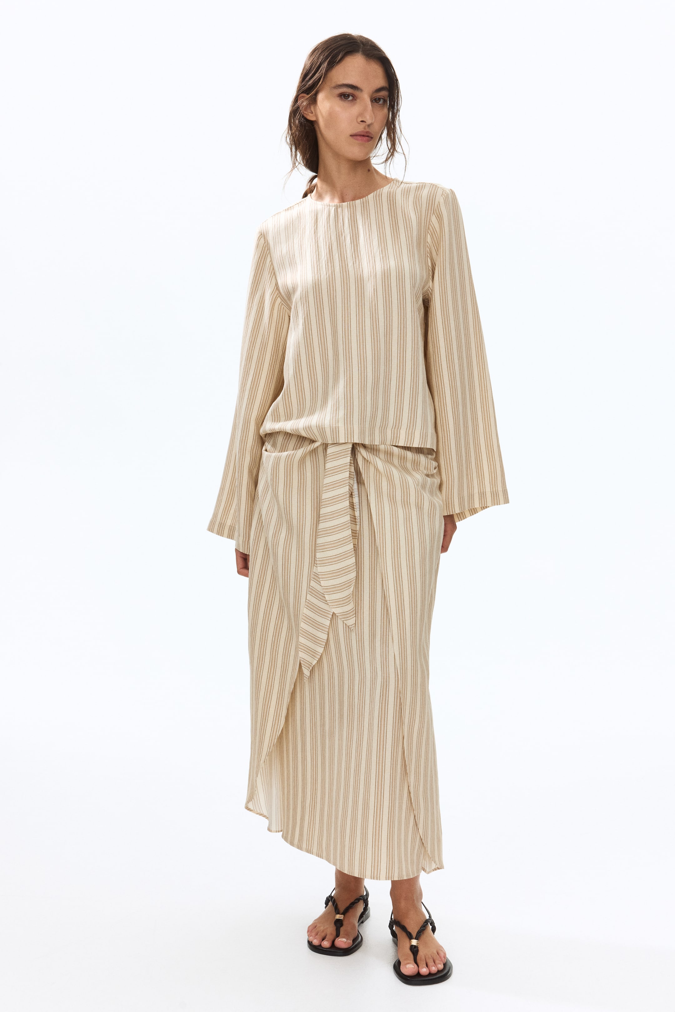 View larger image: Presenting a coordinated two-piece ensemble, comprising a relaxed-fit, long-sleeved top with a round neckline, paired with a midi-length wrap skirt. Both garments exhibit subtle vertical stripes in beige and cream hues. The skirt is accentuated with a front tie and an overlapping panel, adding a touch of elegance.