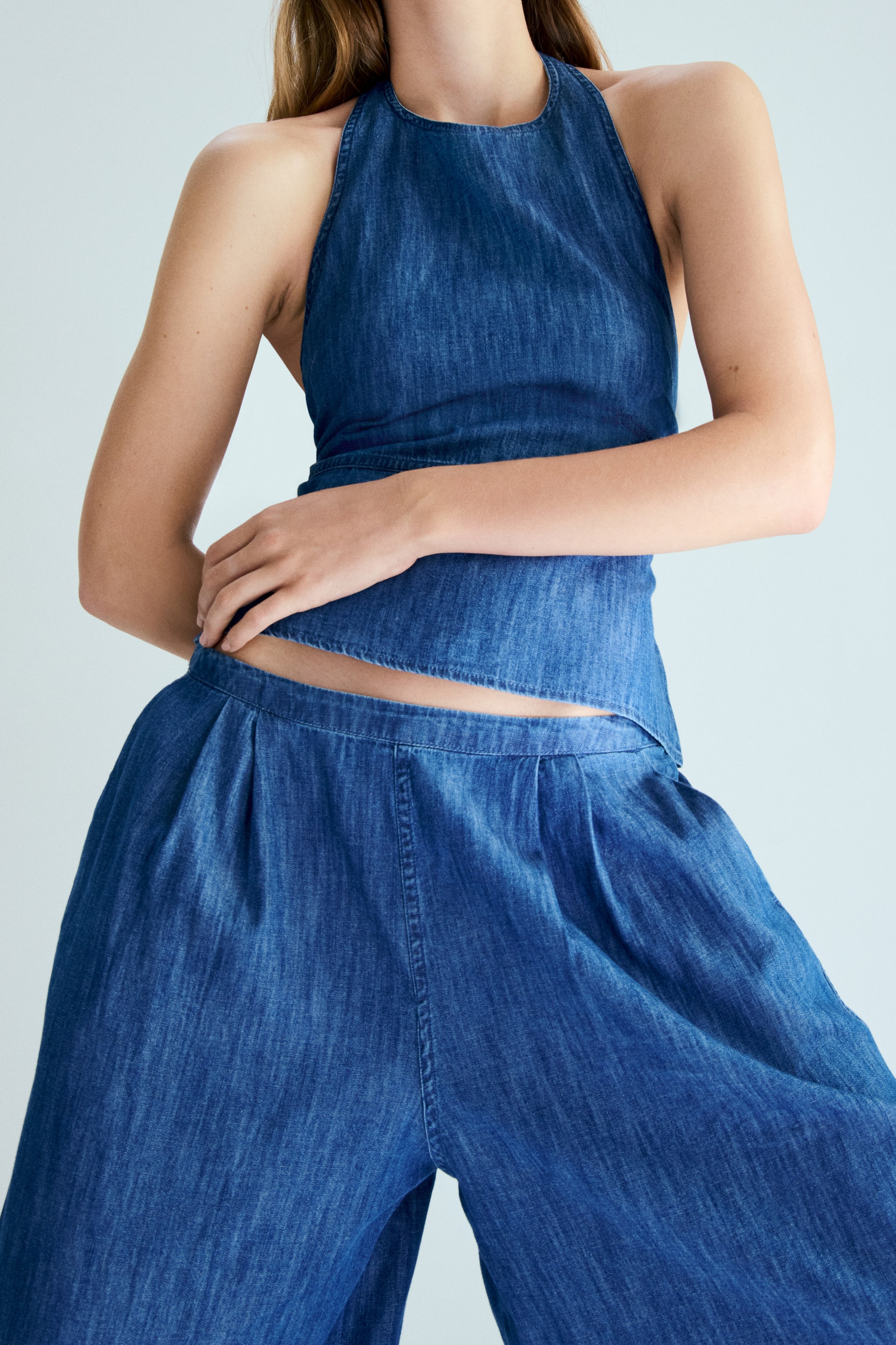 Nagyobb kép megtekintése: Egy kék farmer kétrészes szett, amely egy magas nyakú, nyers szélű, rövidített szegélyű nyakpántos felsőt tartalmaz, valamint hozzá illő magas derekú, bő szárú nadrágot, amely a deréknál ráncokat mutat.