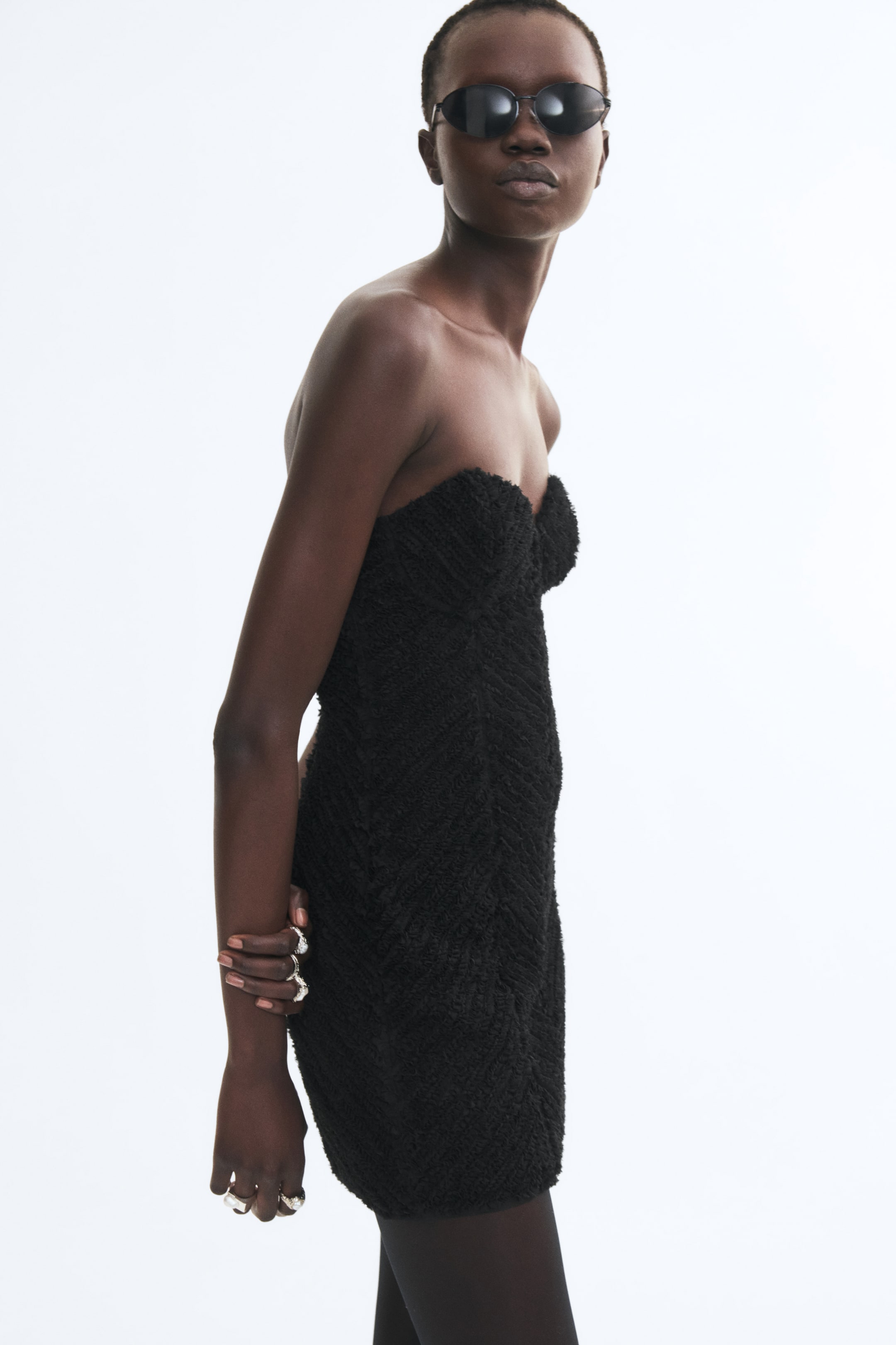 View larger image: A black strapless mini dress with a sweetheart neckline and textured, patterned fabric is being worn by the model. The fitted dress is styled with dark sunglasses, black tights, and silver rings.
