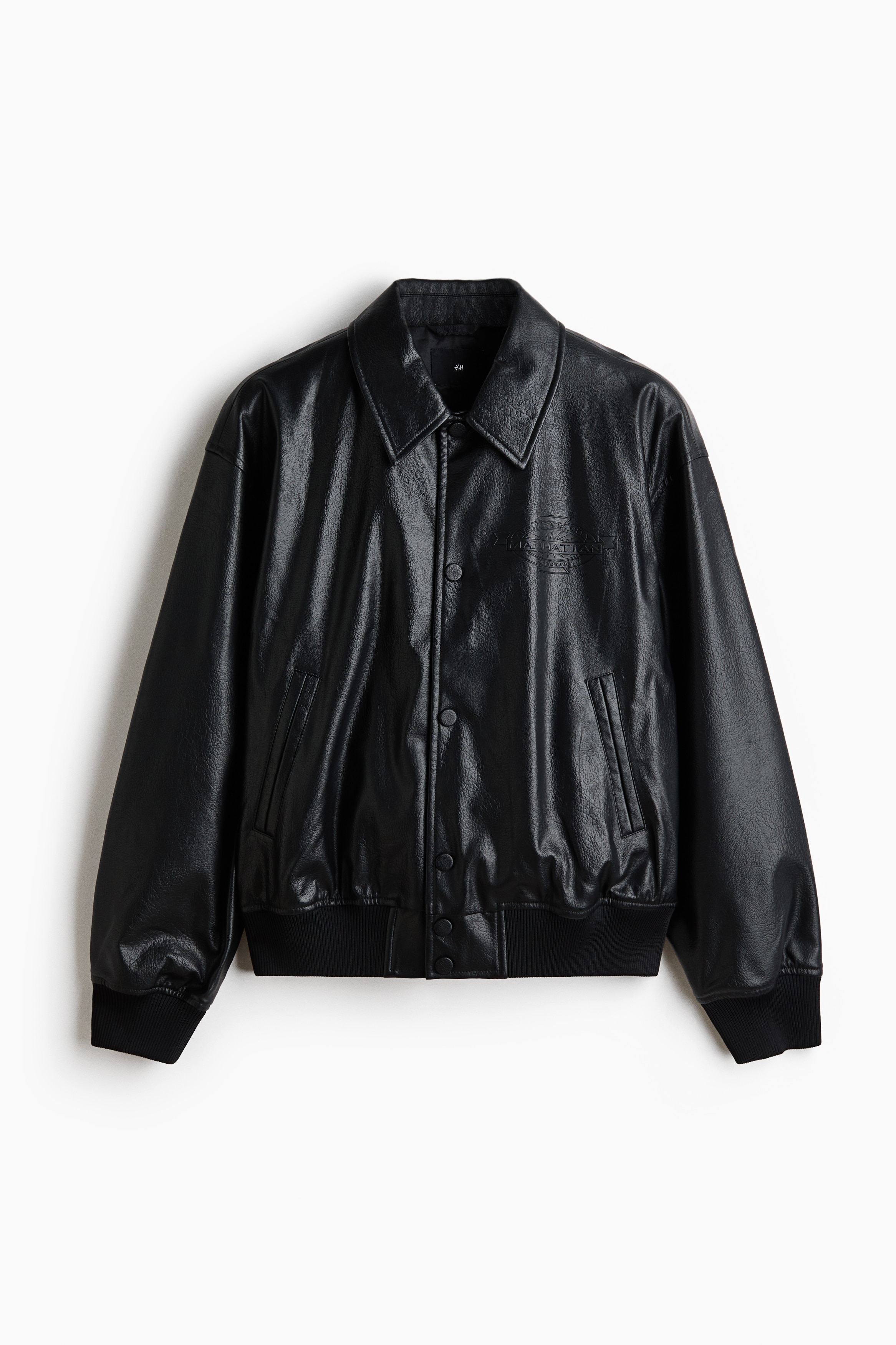 A black jacket in coated fabric is being worn open by the model over a white T-shirt. The jacket features a collar, visible press studs down the front, and wide, ribbed trim at the cuffs and hem.
