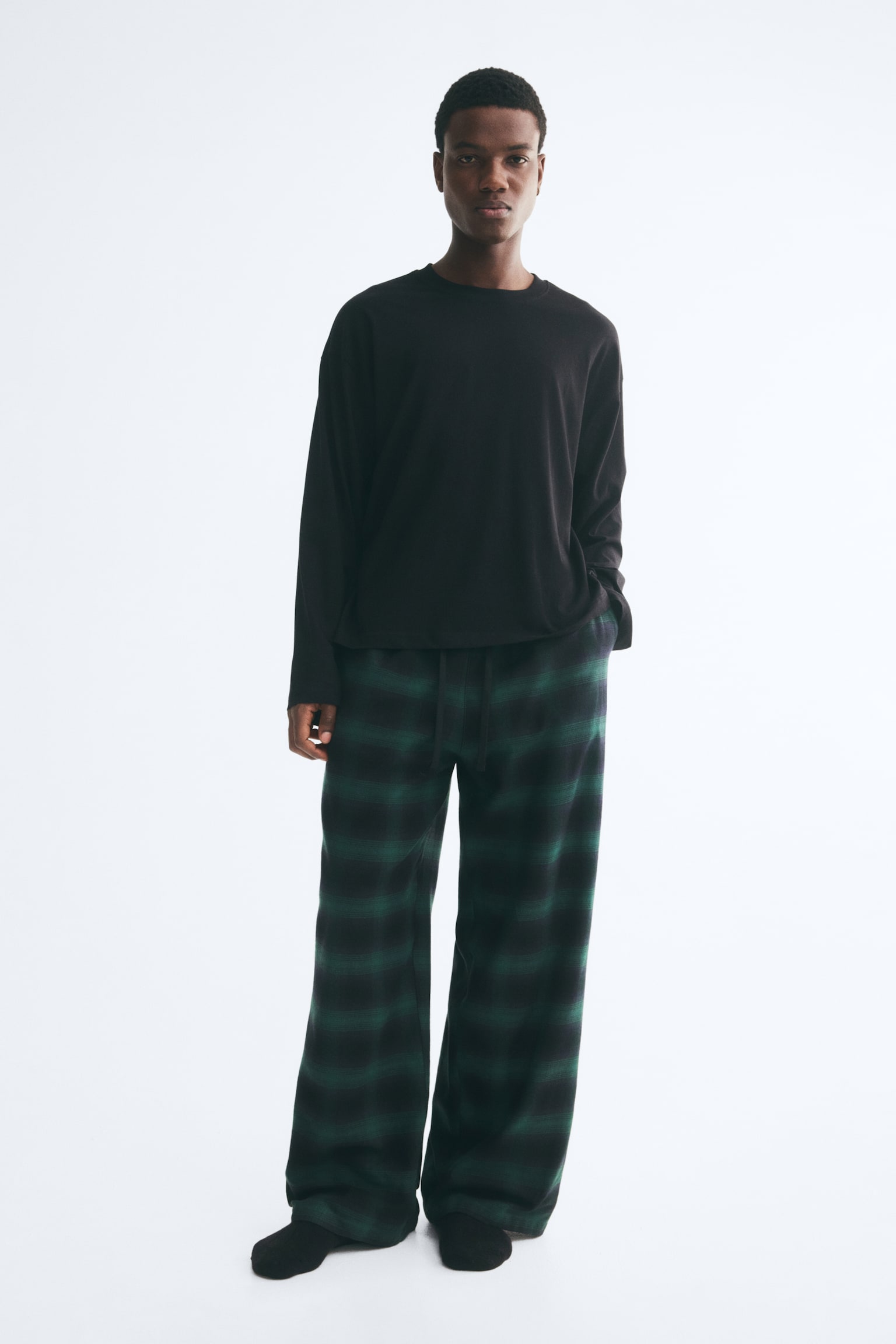 A cotton loungewear set featuring a long-sleeved black jersey top with dropped shoulders and a pair of wide-leg, green and black plaid flannel trousers is being worn by the model. The model is standing with one hand resting inside the trouser pocket, highlighting the relaxed fit and drawstring waist. - 1