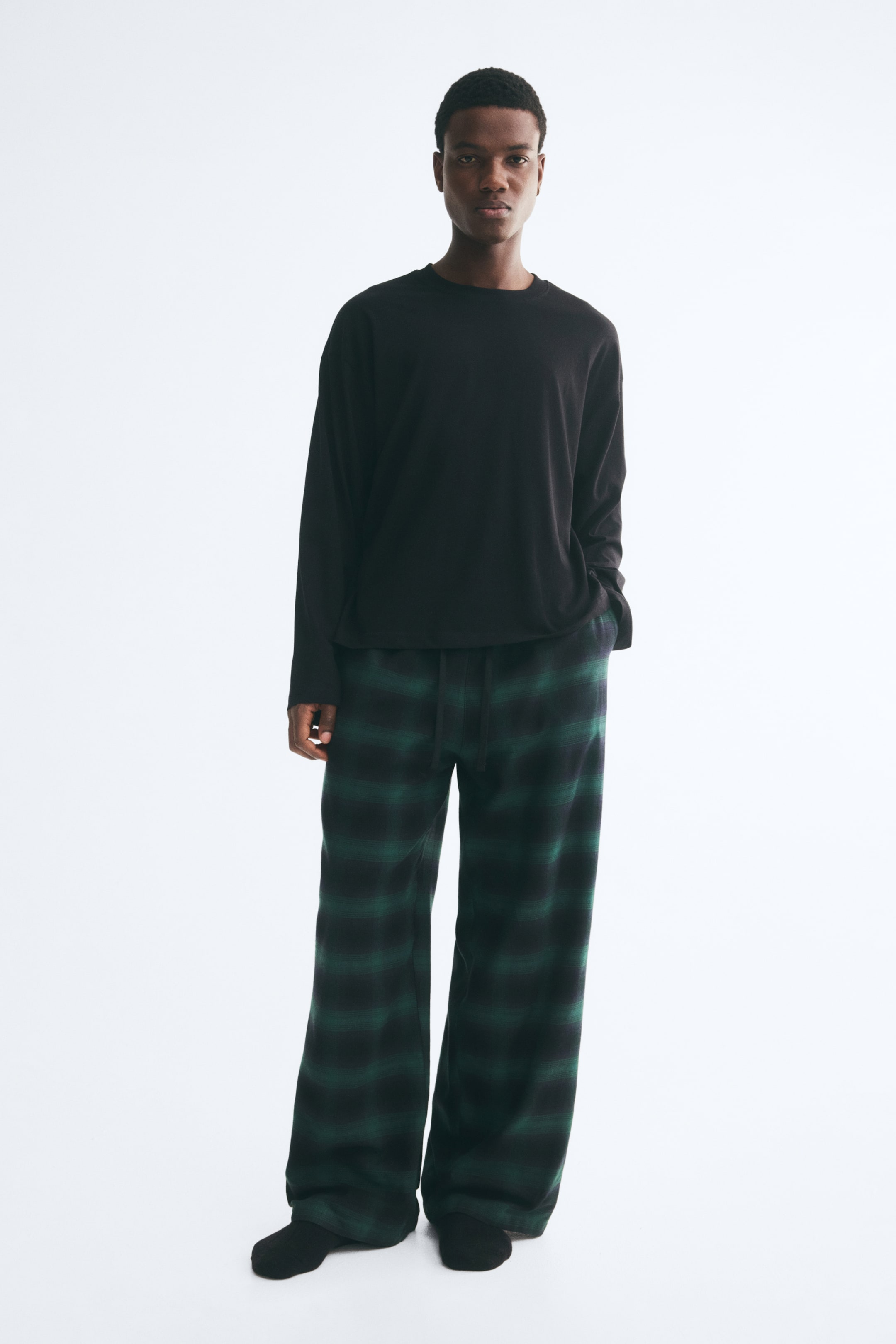 Grotere afbeelding bekijken: Een katoenen loungewear-set bestaande uit een zwarte jersey top met lange mouwen en verlaagde schouders, gecombineerd met een paar wijde flanellen broek in groen en zwart ruitdessin, wordt gedragen door het model. Het model staat met één hand in de broekzak, waardoor de relaxte pasvorm en de taille met trekkoord extra worden benadrukt.