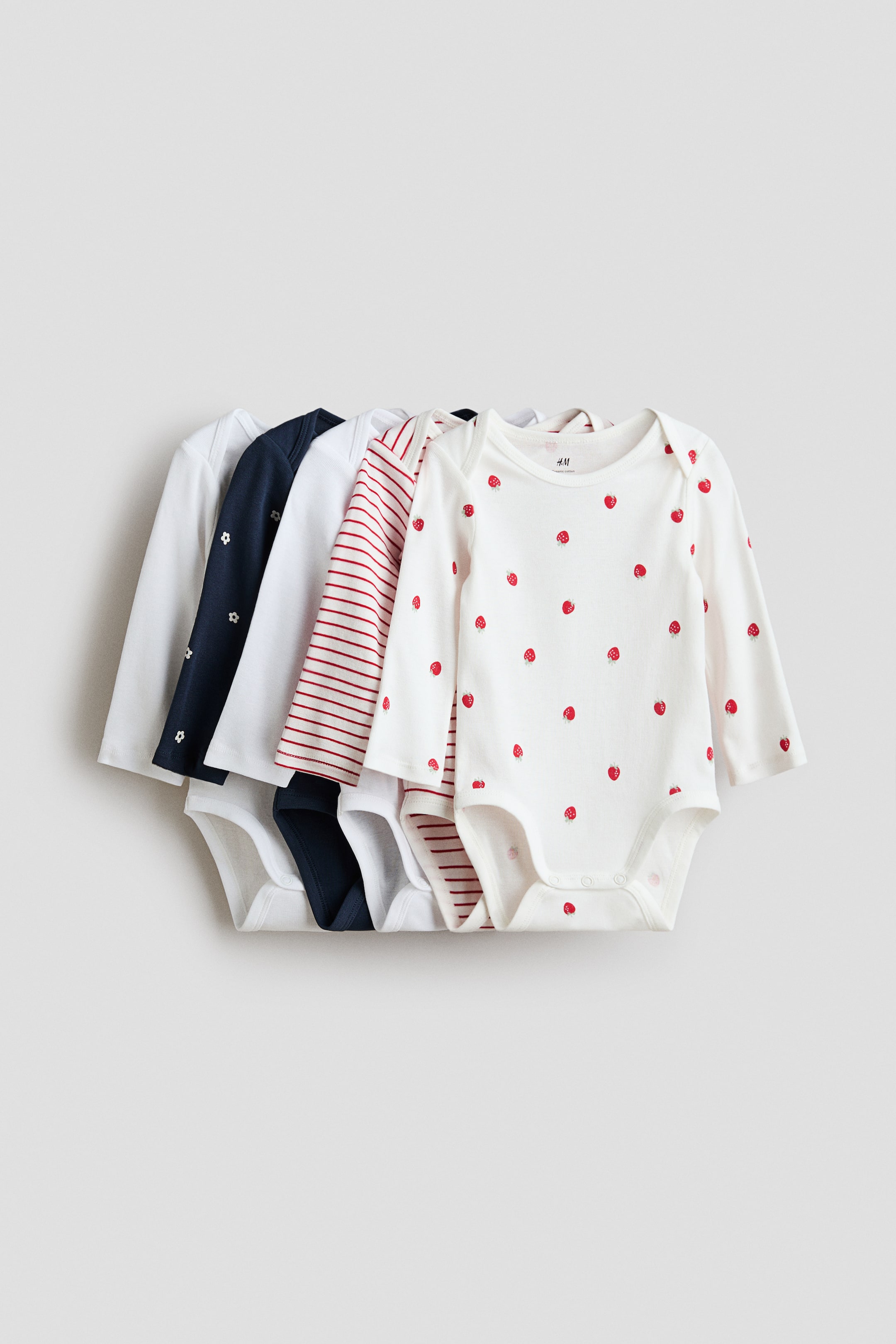 이미지 확대 보기: 부드러운 코튼 저지 소재의 긴소매 보디수트들이 부채꼴로 펼쳐진 다섯 벌 세트로 진열되어 있으며, 각각 겹쳐진(오버랩) 어깨선과 가랑이 부분의 스냅 단추가 특징입니다. 앞쪽 보디수트는 크림색 바탕에 빨간 딸기 패턴이 들어가 있고, 그 뒤로 보이는 보디수트들에는 단색 화이트, 흰 꽃무늬가 있는 네이비, 빨간 줄무늬가 있는 화이트 등이 포함되어 있습니다.
