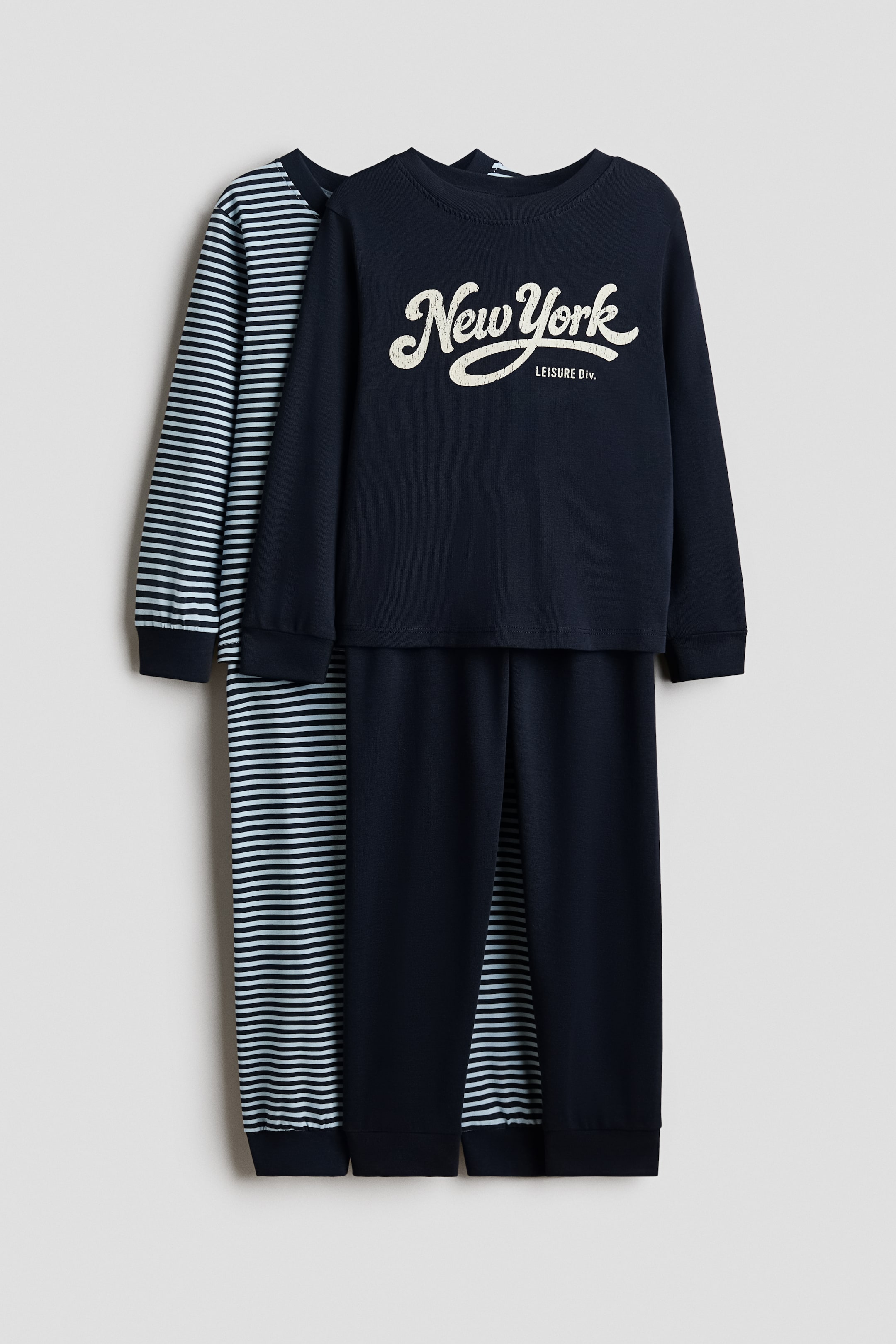 View larger image: Two pyjama sets are shown facing forward, with the front set featuring a dark blue long-sleeved top displaying the text “New York LEISURE DIV.” and matching dark blue trousers with ribbed cuffs. A striped long-sleeved top and matching striped trousers are visible layered behind the first set.
