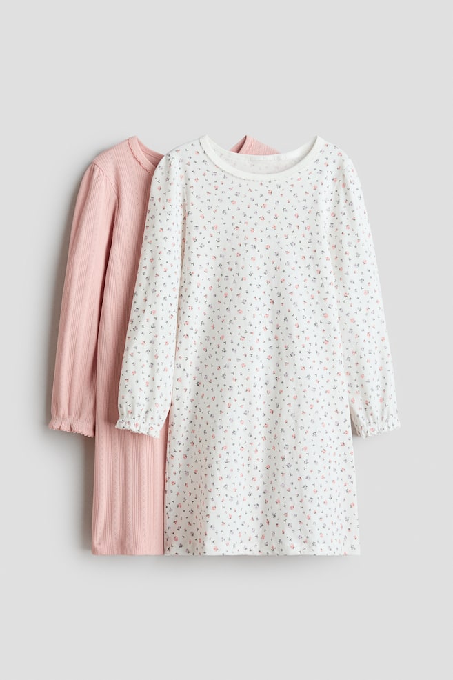 Deux robes à manches longues sont suspendues, face à l'avant. La robe à l'avant est blanche, ornée d'un petit imprimé floral rose et foncé, avec un col rond bordé d'un picot et des poignets froncés. Derrière elle, une robe rose présente une texture côtelée avec des détails ajourés et des poignets volantés. - 1
