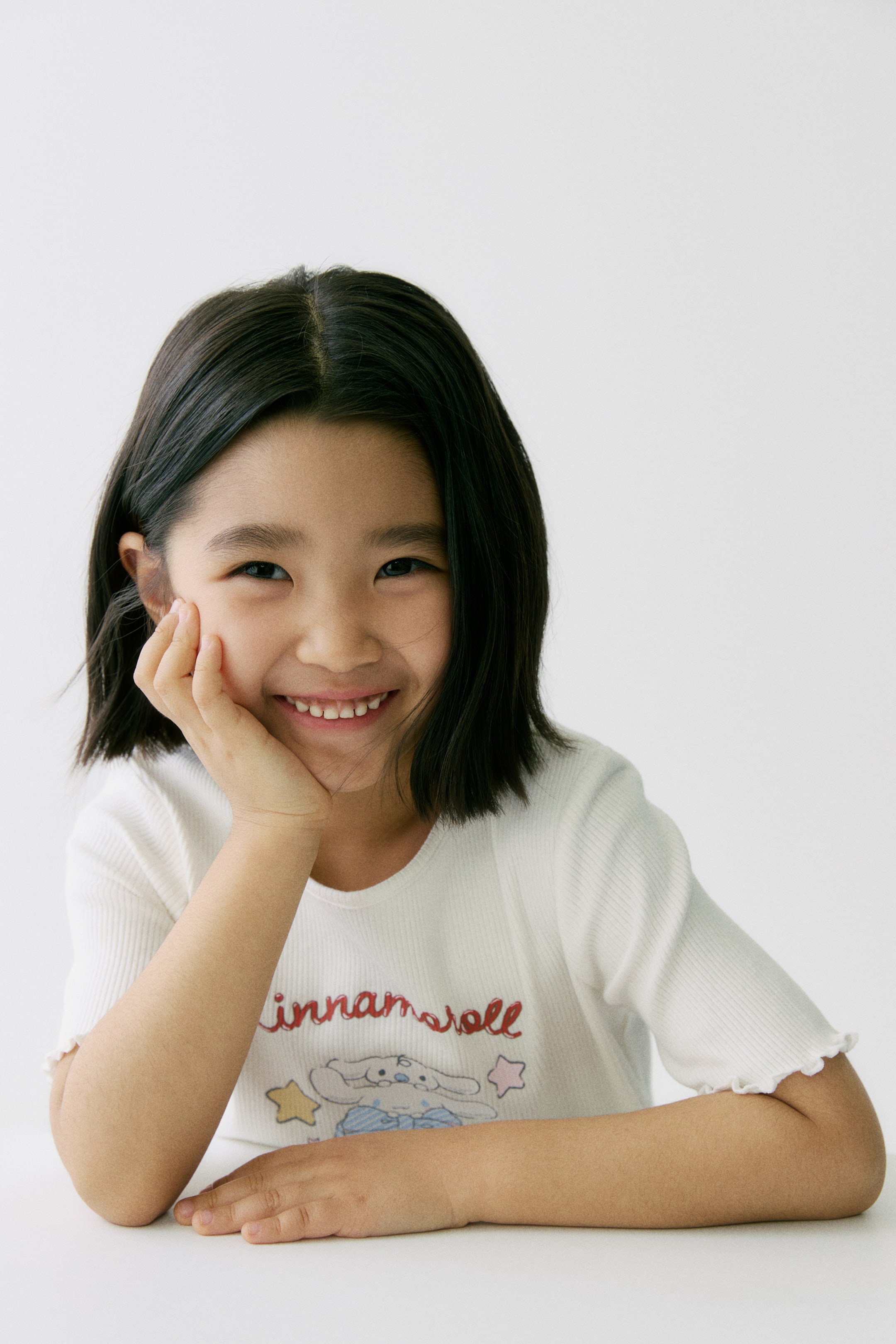 View larger image: A white ribbed cotton jersey T-shirt features a "Cinnamoroll" print motif on the front, a narrow trim around the neckline, and overlocked edges at the short-sleeve cuffs. The model is wearing the T-shirt.