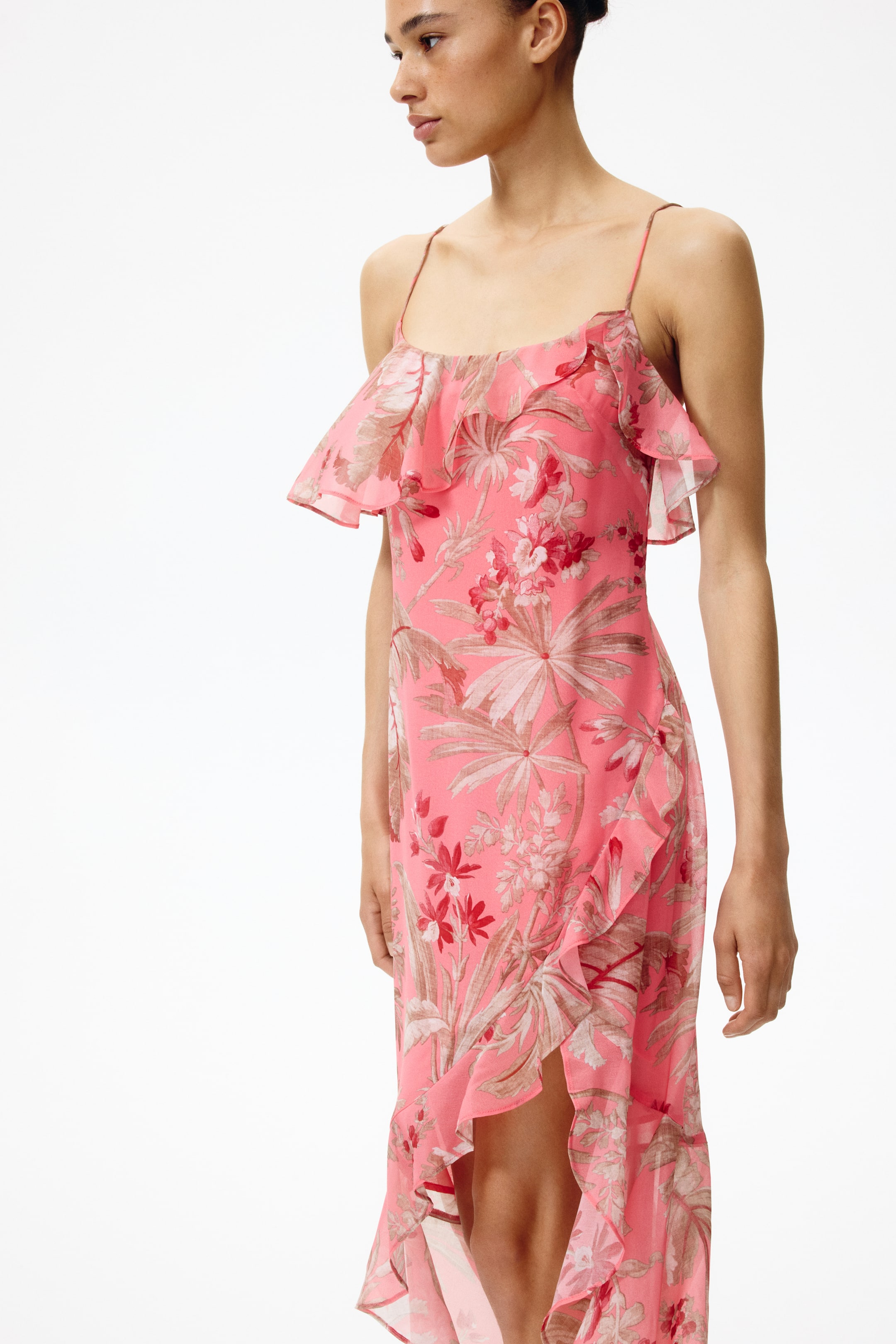 Ver imagen más grande: Vestido con estampado tropical rosa, ligeramente orientado hacia la derecha, que presenta finos tirantes tipo espagueti, un amplio volante en el busto y un dobladillo asimétrico con abertura lateral con volantes.