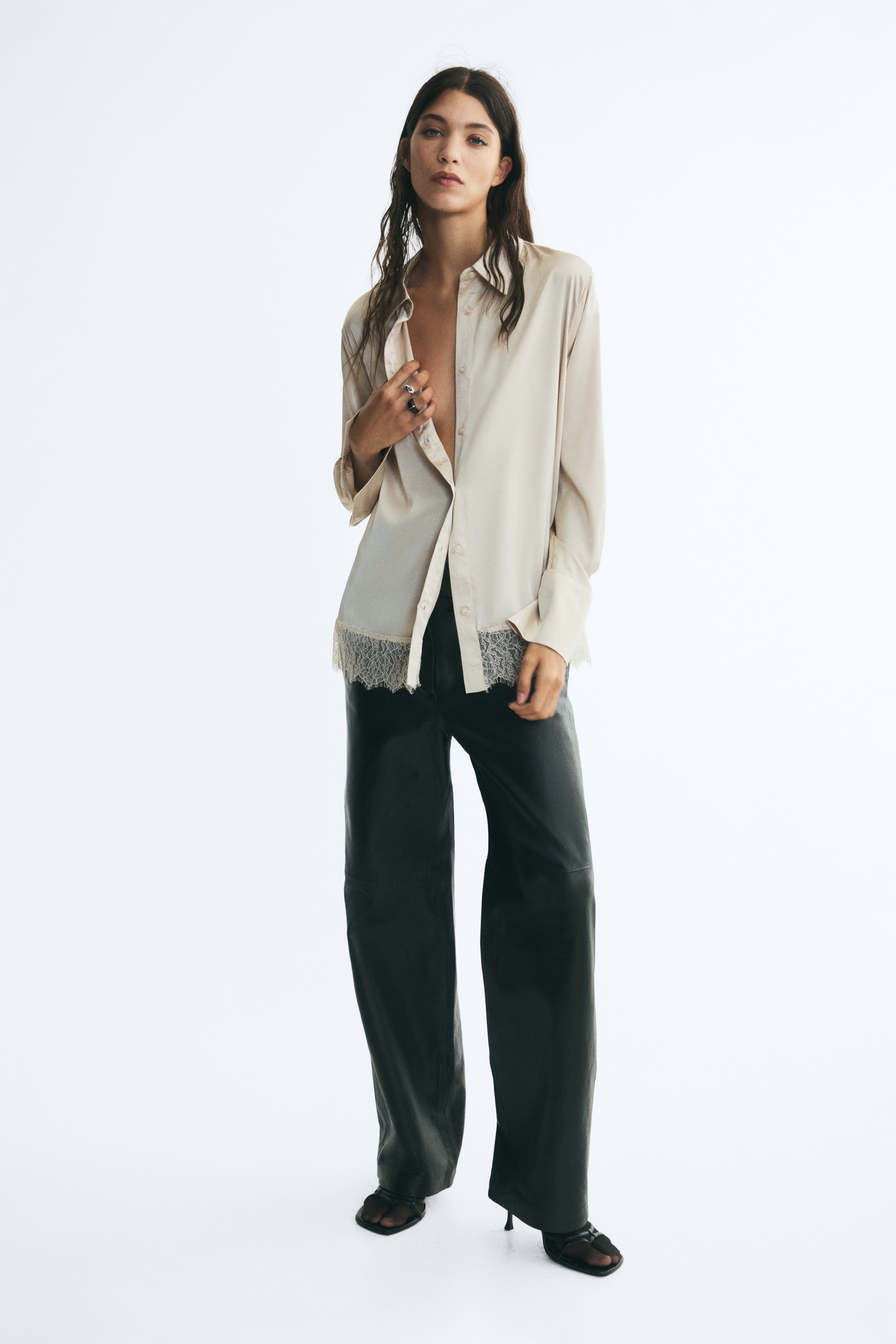 A cream-coloured, long-sleeved satin shirt is shown from the front, featuring a classic point collar, a central button-down placket, and wide cuffs. The straight-cut shirt is detailed with a delicate, semi-sheer floral lace trim along the bottom hem.