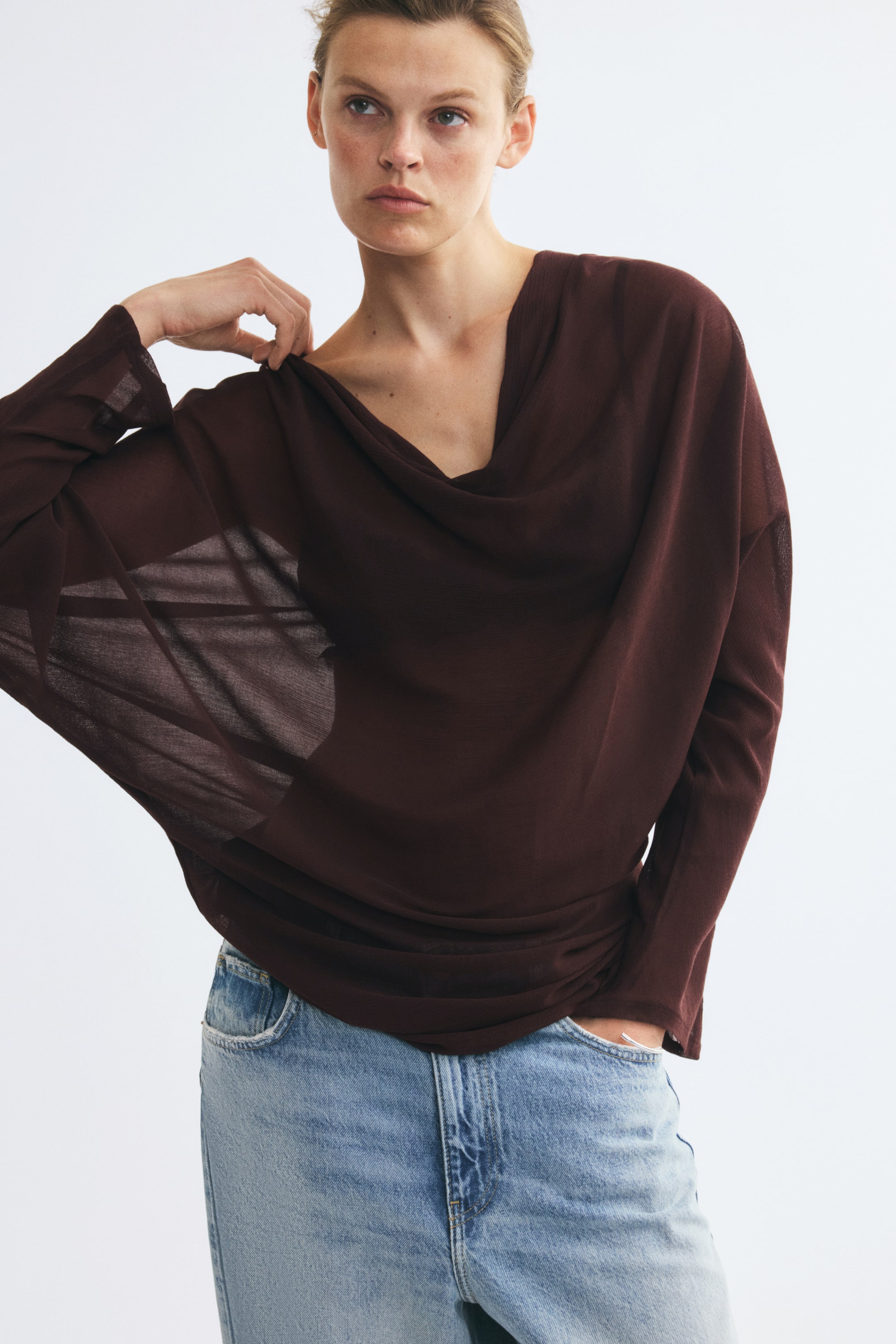 Ampliar la imagen: La modelo lleva puesta una blusa de corte holgado en color café oscuro, tejida en punto fino, mientras ajusta la tela en el hombro con una mano. La blusa presenta un escote drapeado profundo y mangas largas de estilo relajado, y se combina con jeans azul claro.