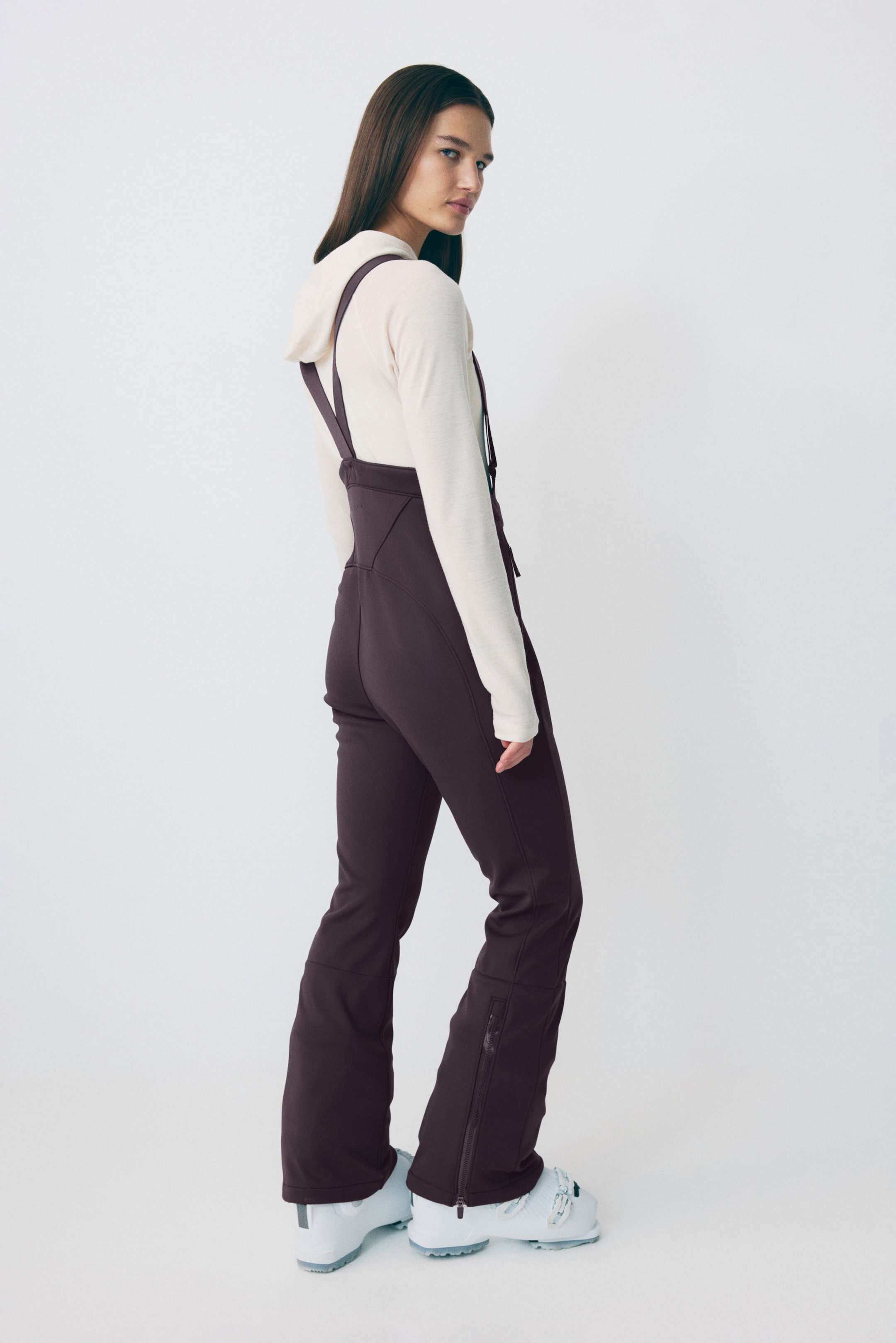 Agrandir l'image: Les pantalons de ski à bretelles de couleur pourpre foncé sont présentés de dos et de côté, avec un haut du corps ajusté et des bretelles. Les pantalons mettent en valeur des jambes évasées avec des fermetures éclair sur le bas extérieur de la jambe, et sont portés par-dessus des bottes de ski avec un haut à manches longues de couleur claire visible en dessous.