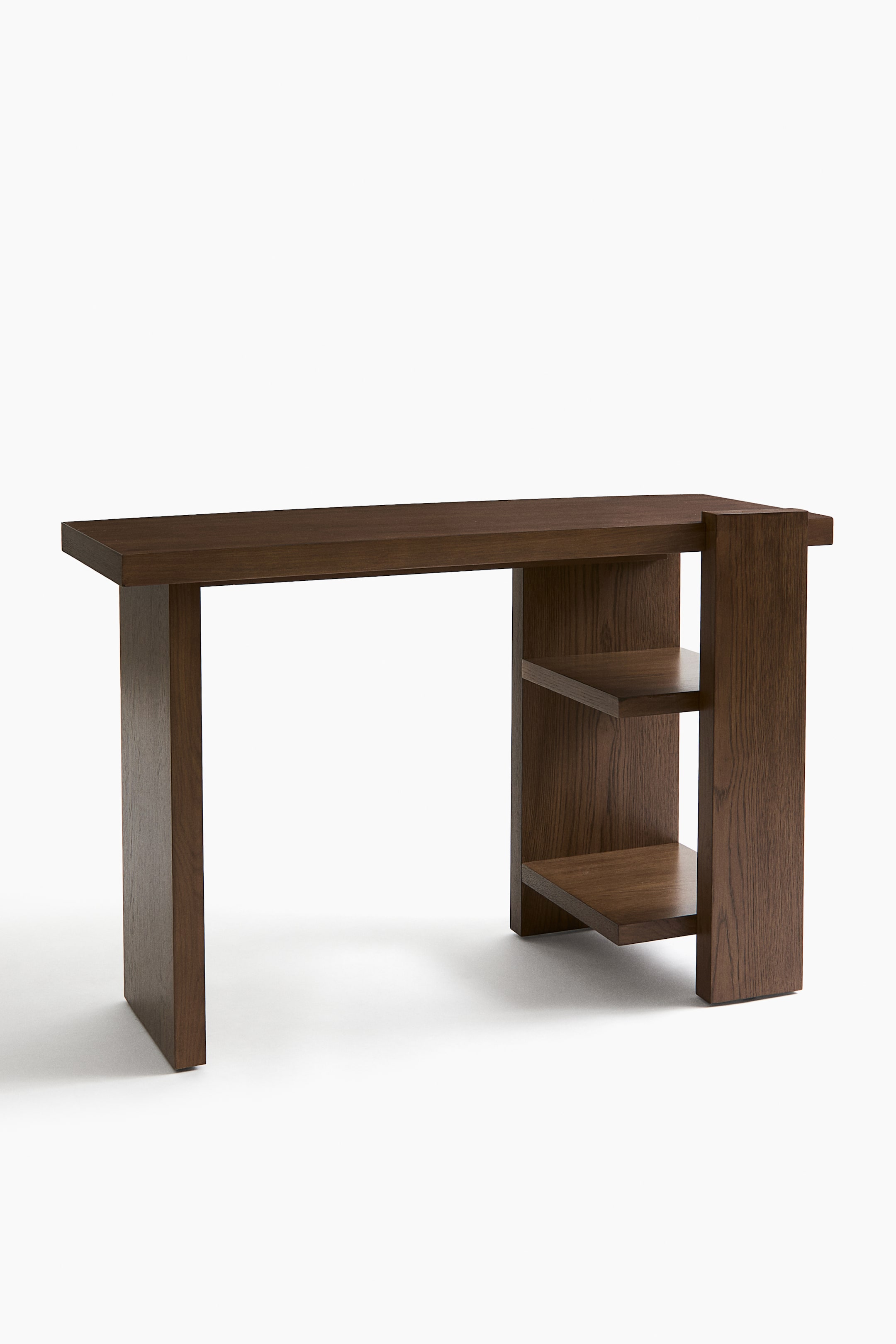 Visualizza l'immagine ingrandita: Un tavolo in legno marrone scuro mostra una ricca venatura del legno sulla superficie rettangolare del piano, ripreso da una leggera prospettiva frontale inclinata. Presenta una gamba a pannello pieno sul lato sinistro e un’unità di scaffalatura integrata sul lato destro, composta da due ripiani aperti.