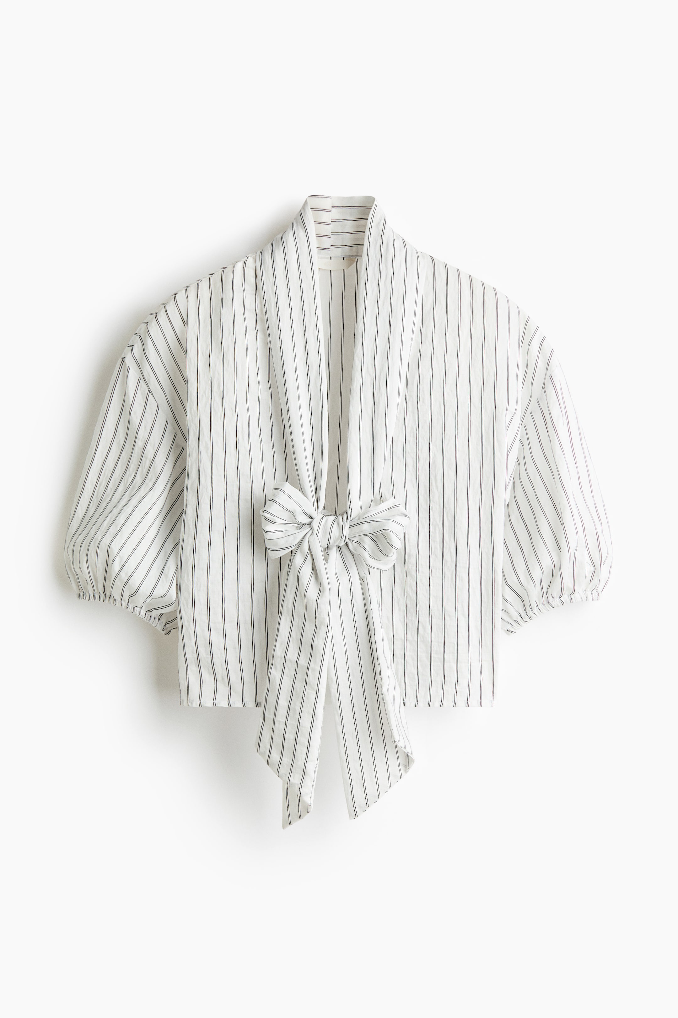 Agrandir l'image: Une blouse courte à coupe décontractée en tissu blanc froissé à rayures verticales foncées est présentée de face, avec un profond col en V et un col lavallière marqué noué au centre. La blouse met en valeur des épaules tombantes et de courtes manches bouffantes volumineuses, terminées par des poignets élastiqués.