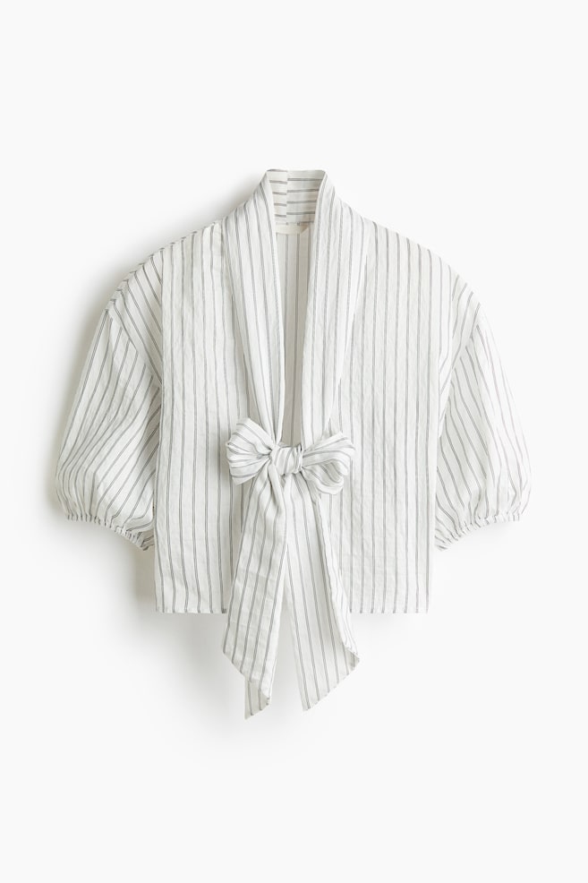 Una blusa corta dalla vestibilità rilassata, in tessuto bianco stropicciato con sottili strisce verticali scure, presentata in una vista frontale, con profonda scollatura a V e un importante colletto con fiocco annodato al centro. La blusa mette in evidenza spalle scese e maniche corte a palloncino voluminose, rifinite con polsini elasticizzati. - 1
