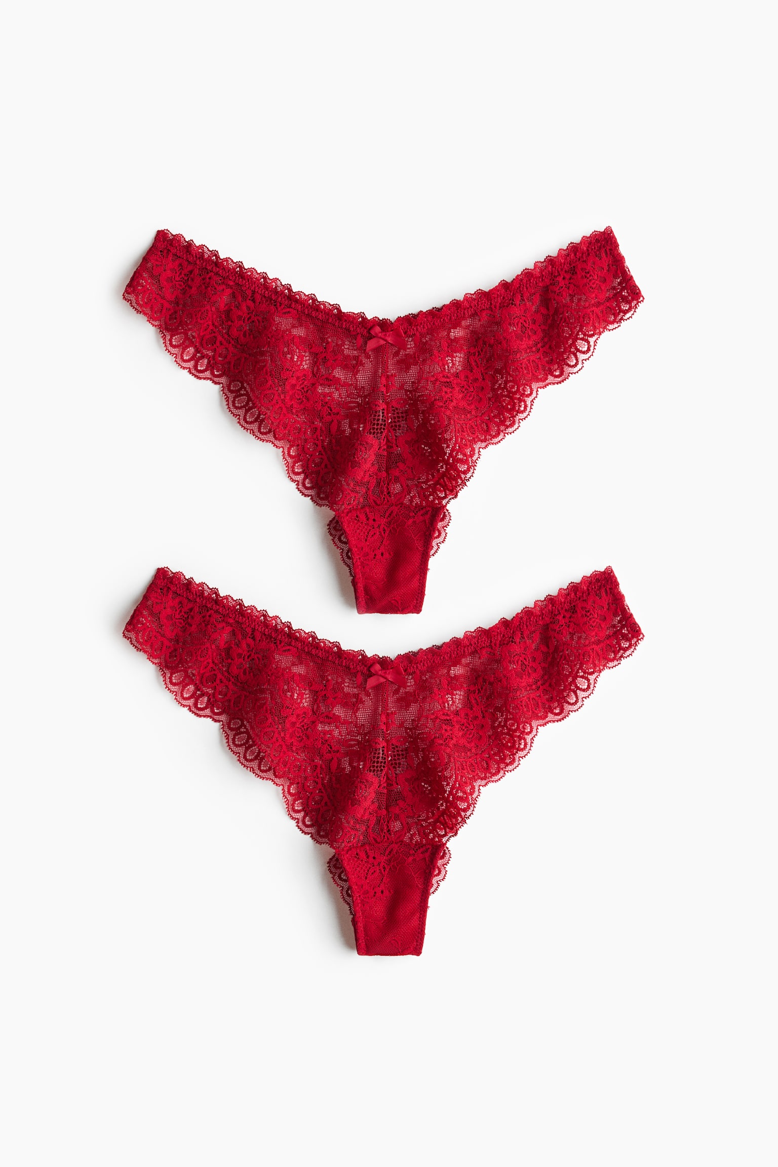 Two pairs of red lace briefs are shown from the front, featuring a low-rise, V-shaped waistband with a small decorative bow at the centre. The briefs showcase a high-cut design made from sheer floral lace with scalloped edges.