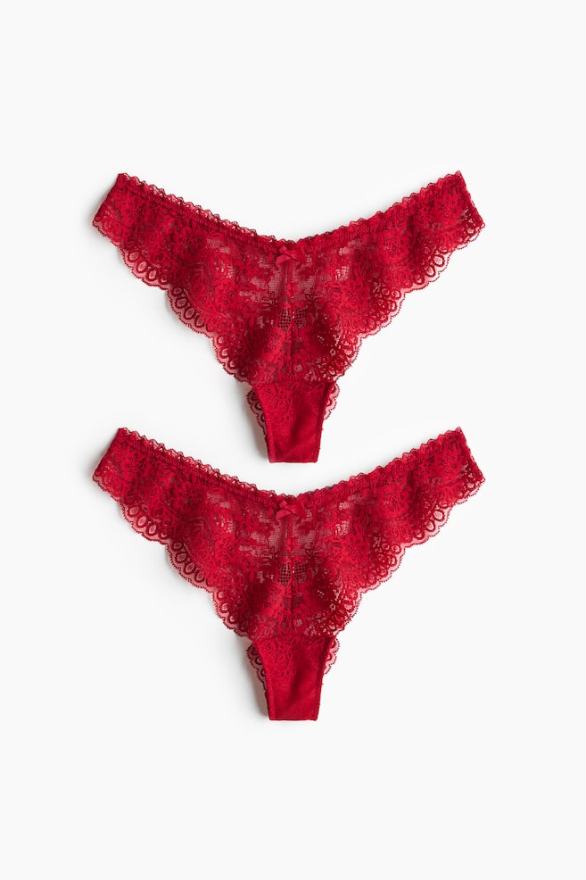 Two pairs of red lace briefs are shown from the front, featuring a low-rise, V-shaped waistband with a small decorative bow at the centre. The briefs showcase a high-cut design made from sheer floral lace with scalloped edges. - 1