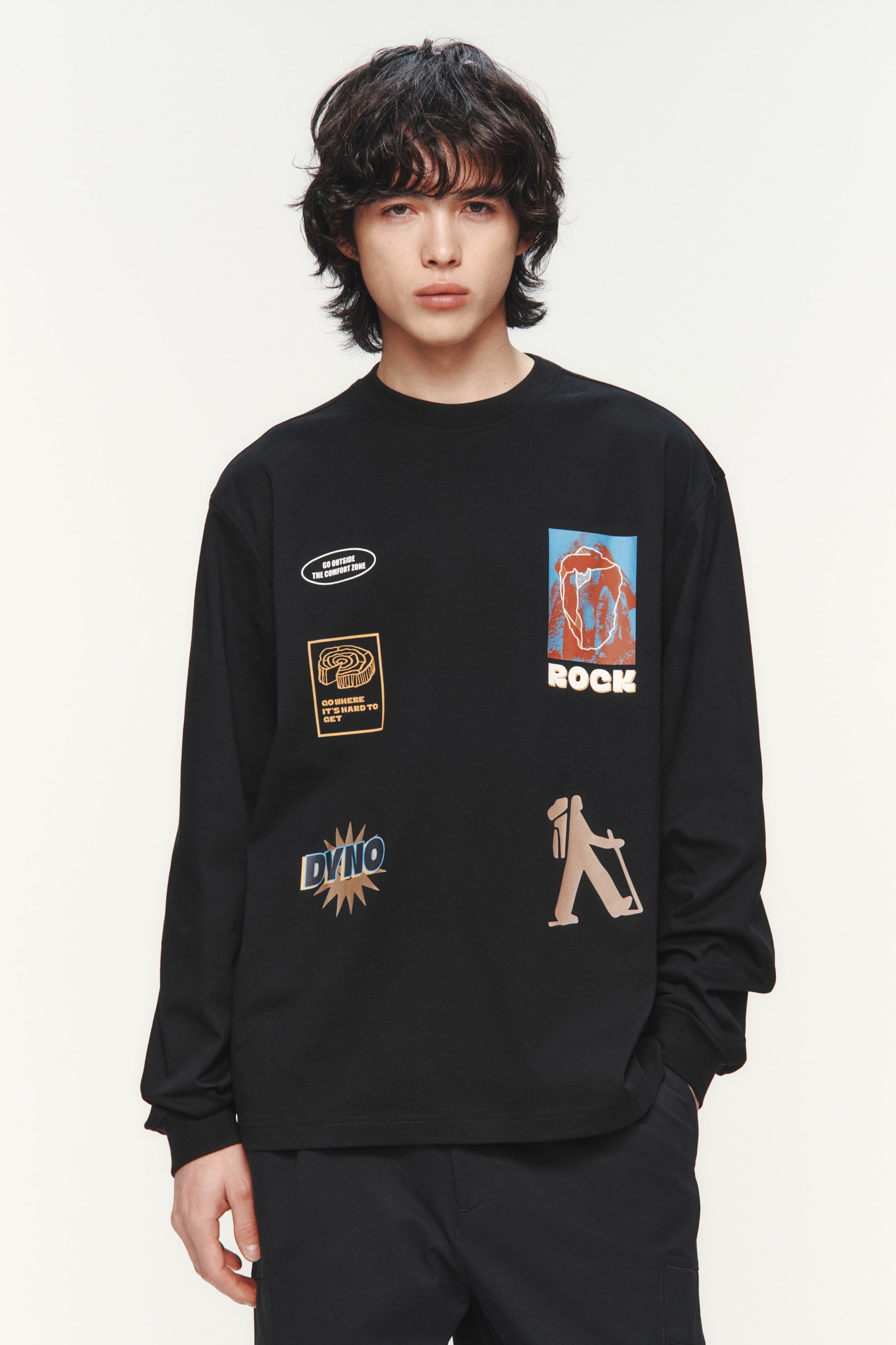 拡大画像を表示: 正面から撮影された黒の長袖クルーネックTシャツには、プレーンなカフスが施され、複数の個性的なグラフィックが特徴です。楕円形の「GO OUTSIDE THE COMFORT ZONE」パッチ、渦巻き状の「GO WHERE IT'S HARD TO GET」パッチ、「DYNO」テキストグラフィック、抽象的な岩の形の下にある「ROCK」テキストグラフィック、バックパックを背負ったスタイリッシュな人物が描かれています。